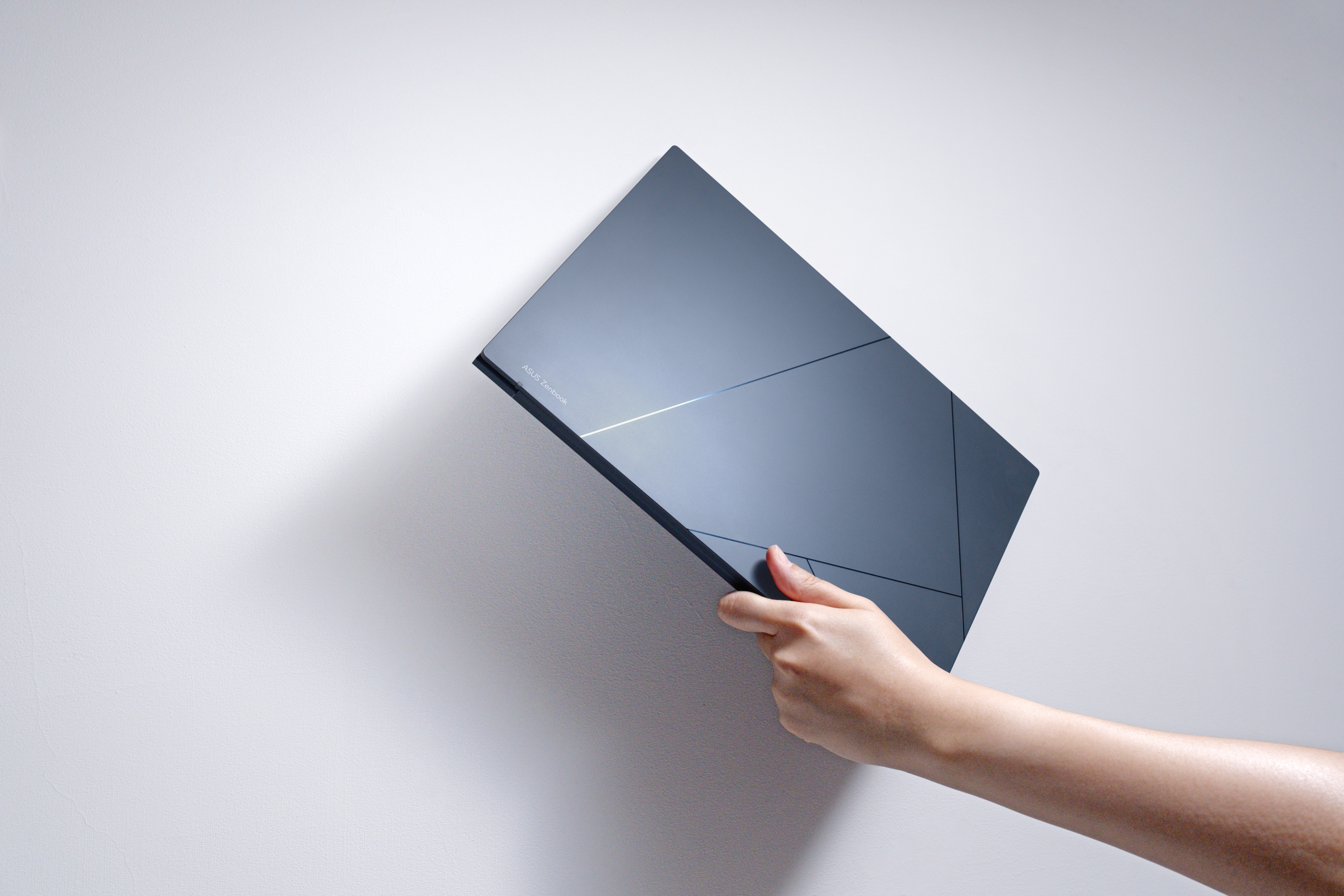 Een hand houdt een donkergrijze laptop vast tegen een witte muur. De laptop heeft een geometrisch ontwerp. Het merk is ASUS Zenbook.