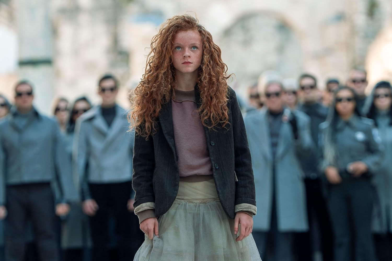 Ein junges Mädchen mit roten Locken steht vor einer Menschenmenge. Sie trägt eine dunkle Jacke und einen hellen Rock. Dahinter tragen Leute Sonnenbrillen.