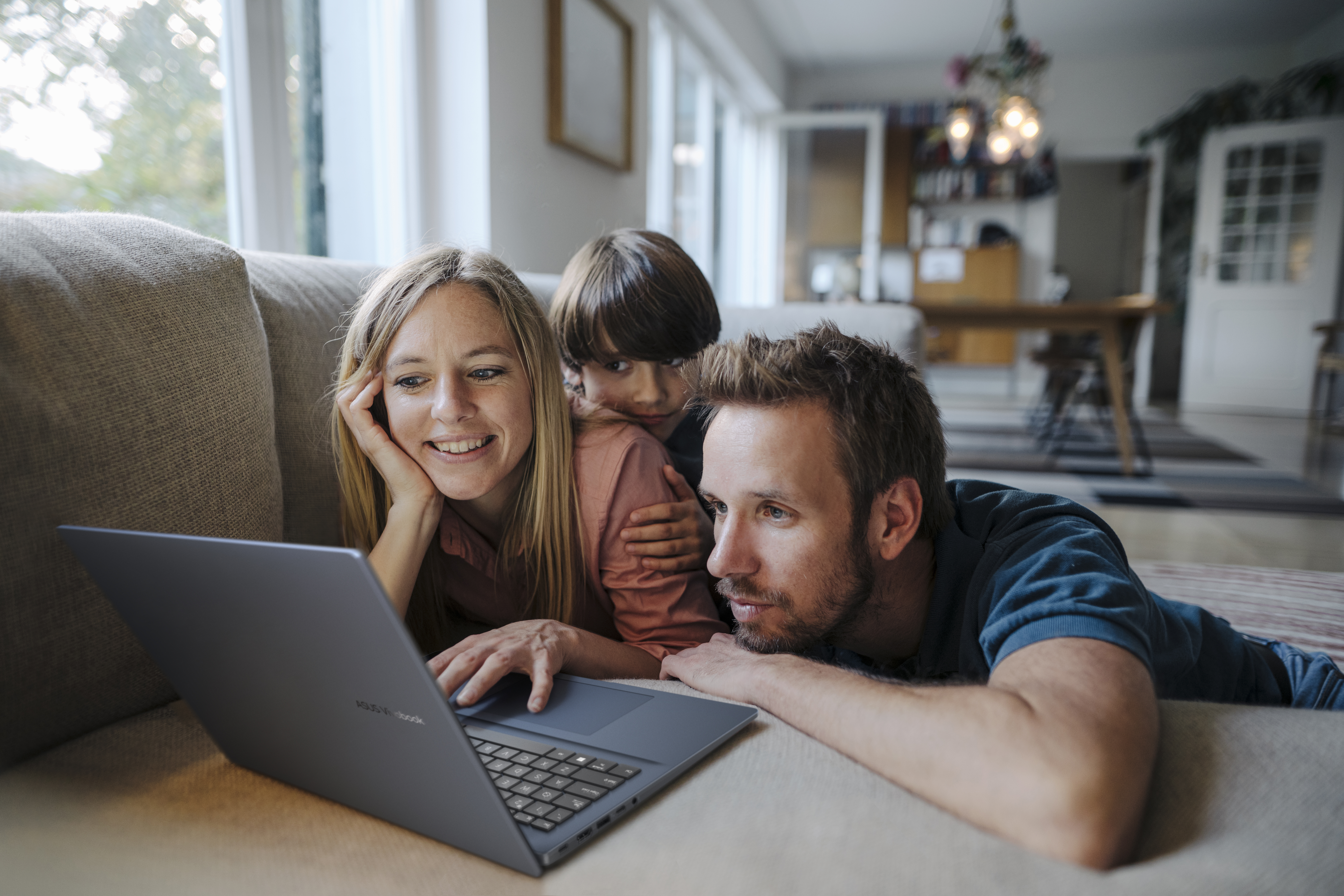 A family of three looks at a laptop. The woman smiles, the man looks intently. A child hugs them. They are on a sofa.