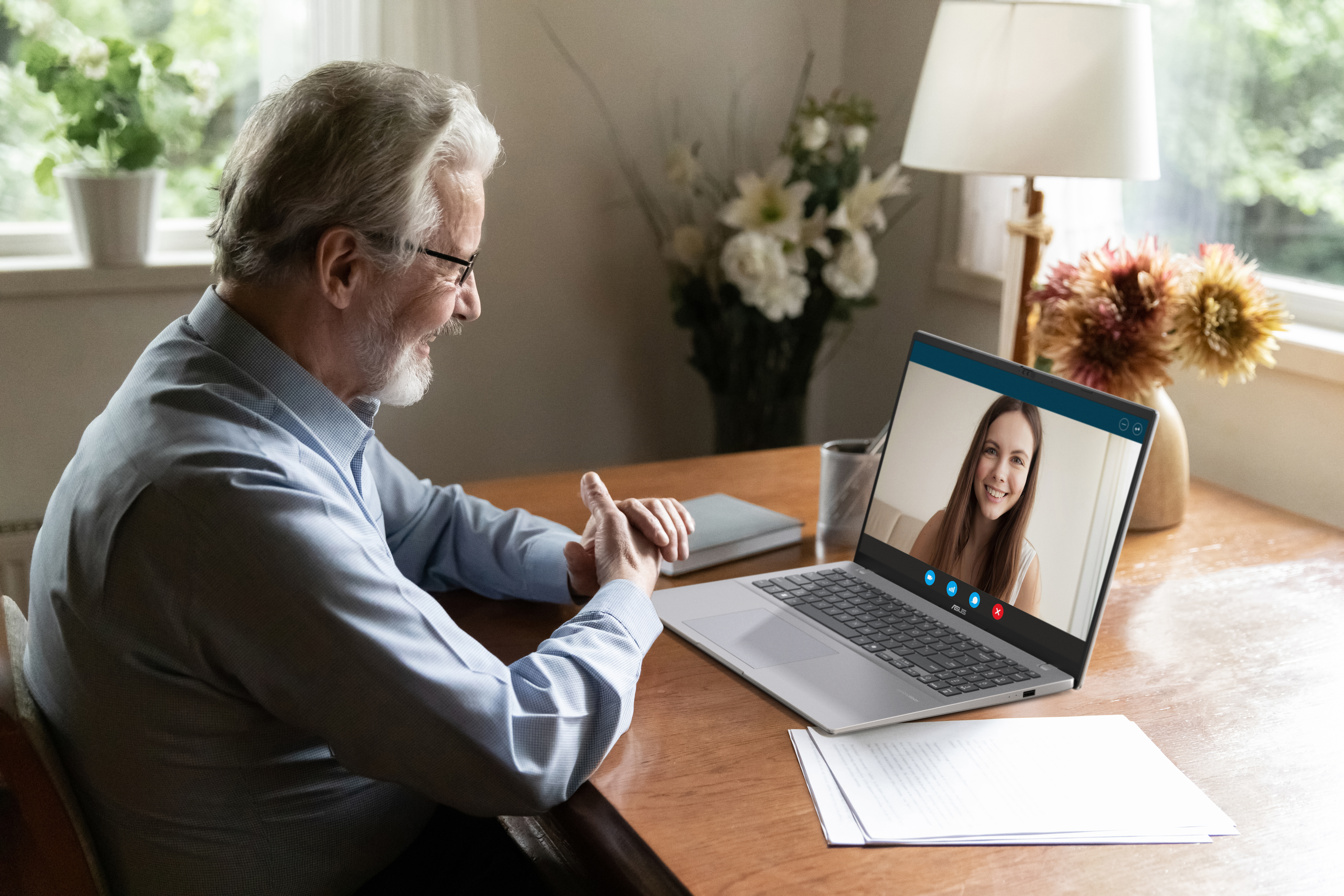 An elderly man smiles at a laptop screen displaying a woman. Flowers and a lamp are visible in the background.