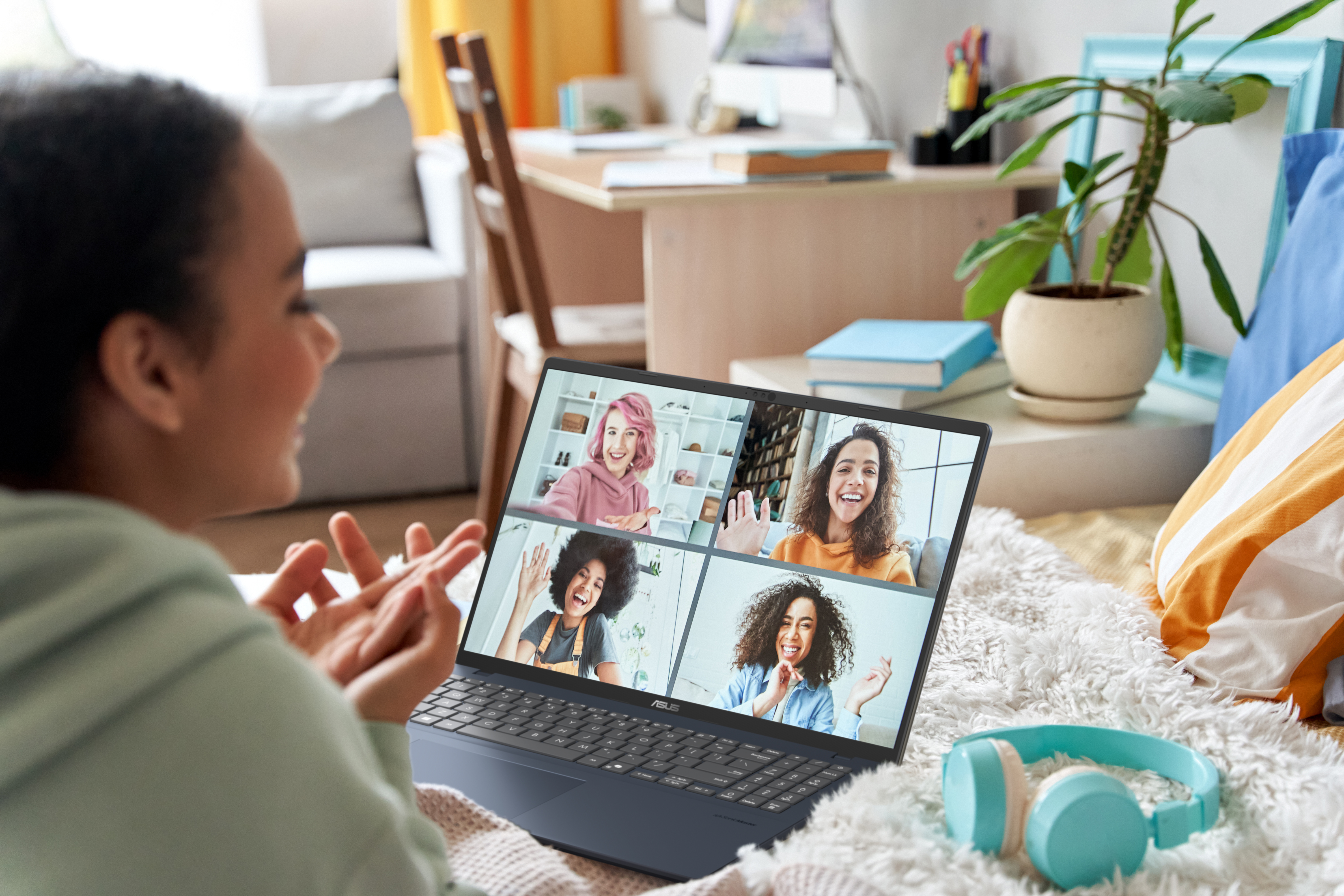A woman on a video call with 4 others on a laptop. A pair of headphones and a pillow are in the foreground.