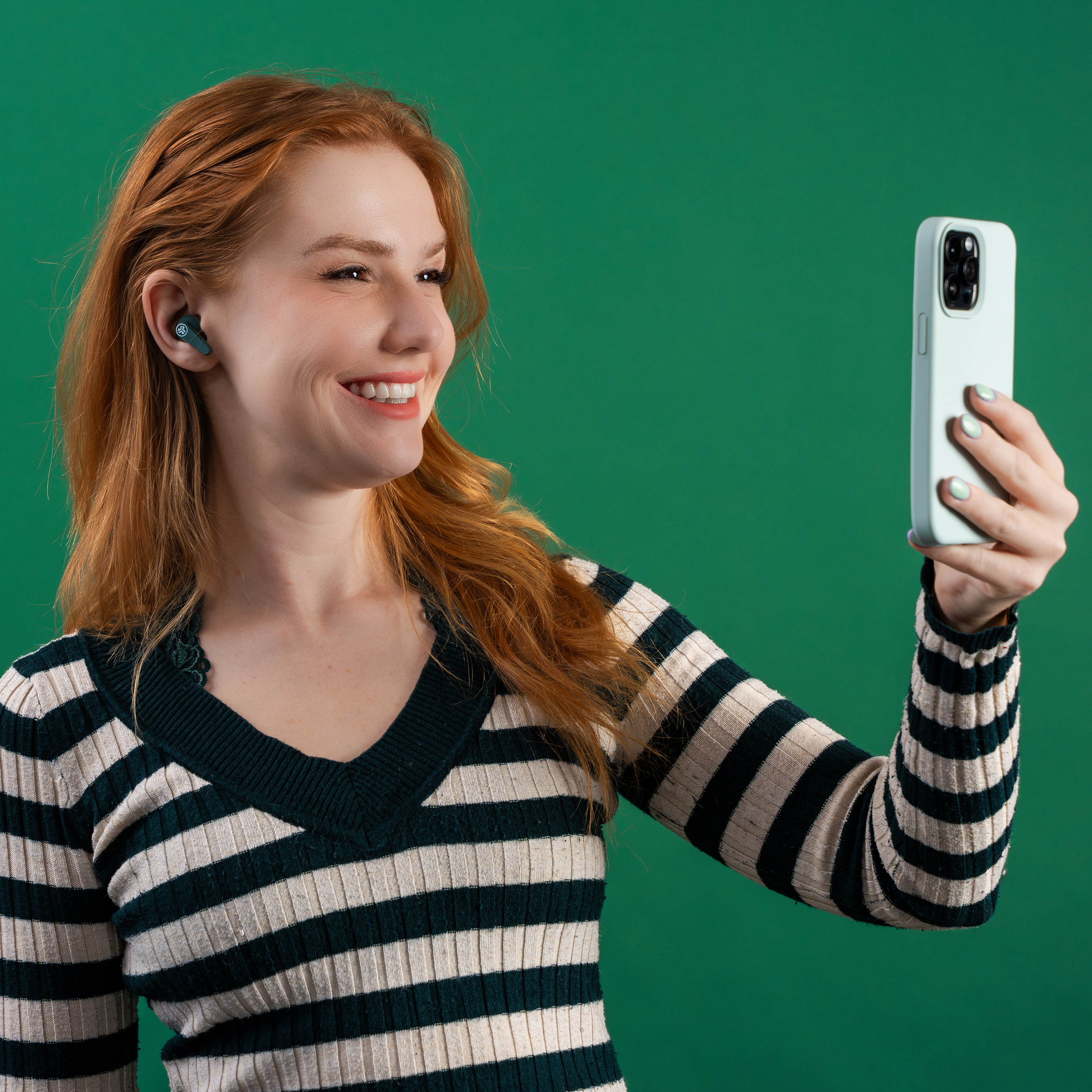 Eine rothaarige Frau lächelt beim Selfie-Machen. Sie trägt Ohrhörer und ein gestreiftes Hemd.