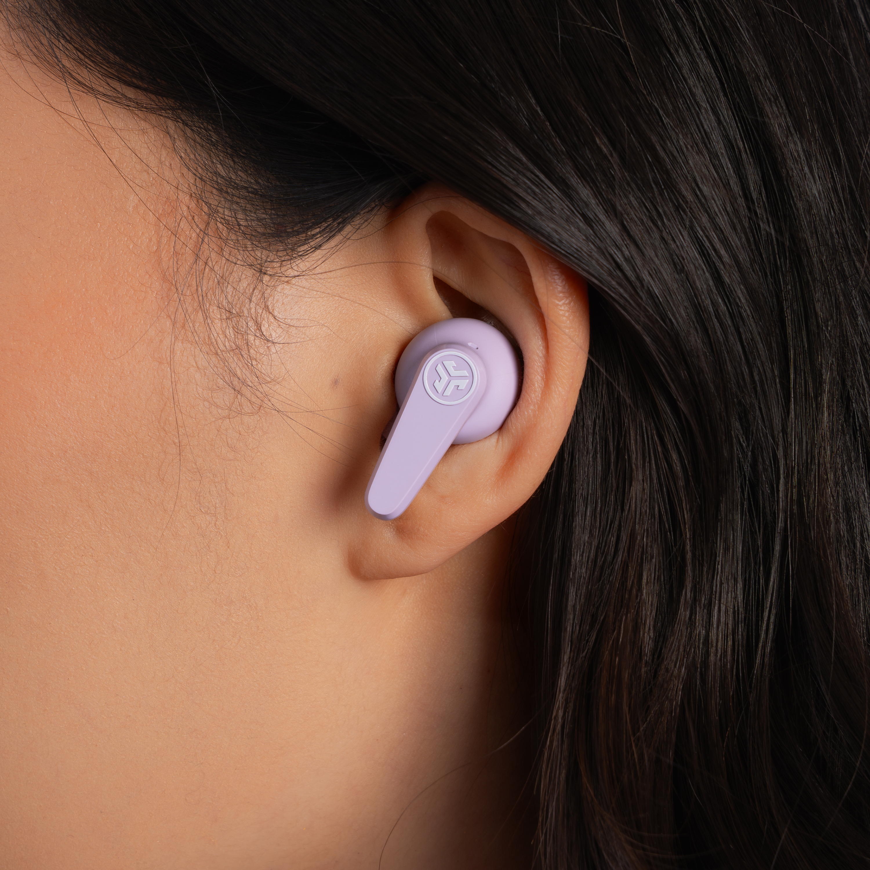 Ein Ohr einer Person mit einem violetten Ohrhörer. Dunkles Haar umgibt das Ohr, Haut ist sichtbar.
