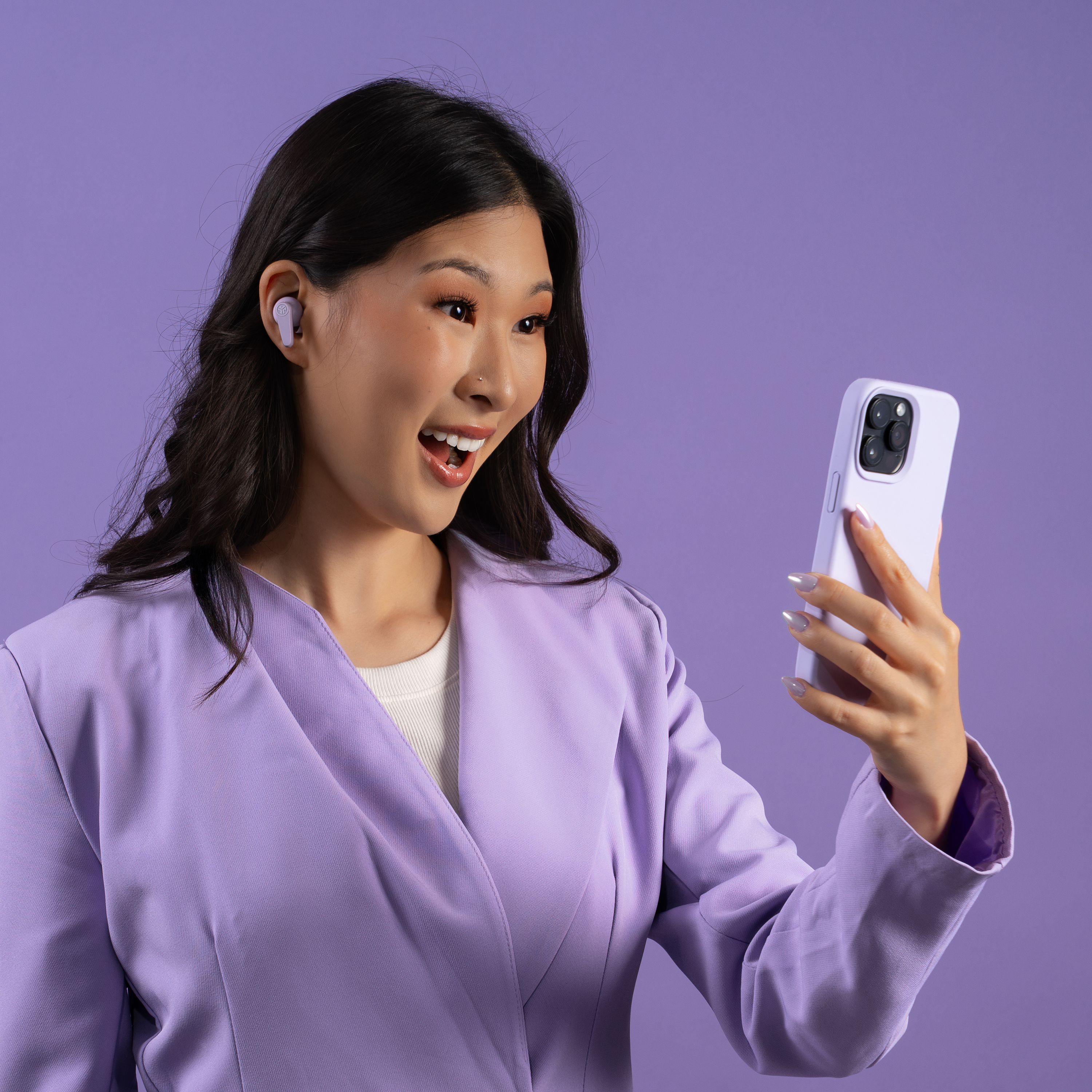 Frau in lila Blazer, benutzt ein Telefon, trägt Ohrhörer. Lila Hintergrund. Sie lächelt.