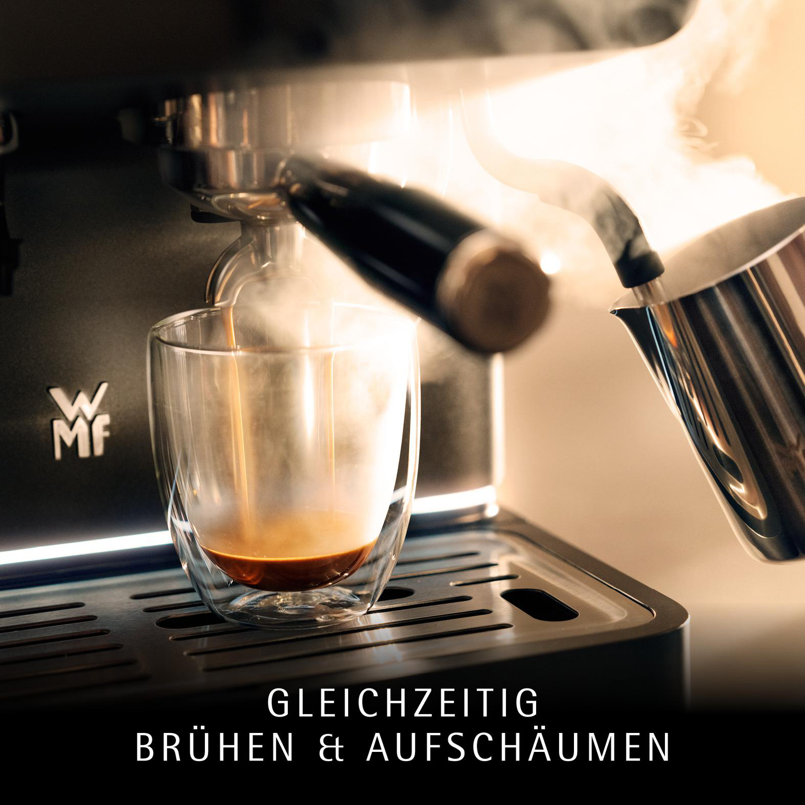 Eine Espressomaschine brüht Kaffee in ein Glas. Dampf steigt auf. Ein Milchaufschäumer gießt Milch.