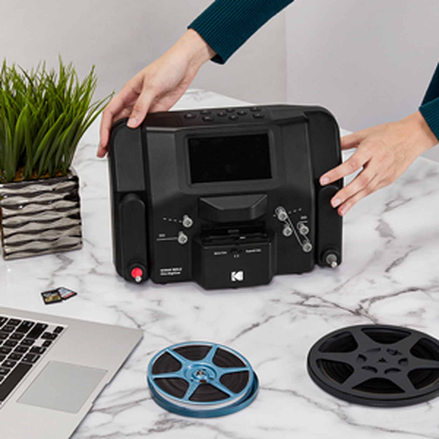 Le mani tengono uno scanner per film nero. Un laptop, bobine di film e una pianta sono su una superficie di marmo. Una scheda di memoria è accanto al laptop.