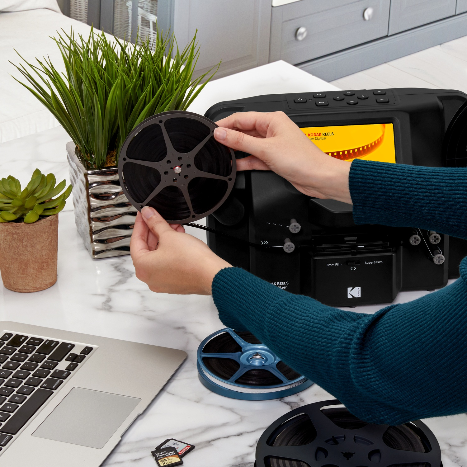Una persona tiene in mano una bobina di film vicino a un digitalizzatore di film. Un laptop, piante e schede SD si trovano sulla superficie di marmo.