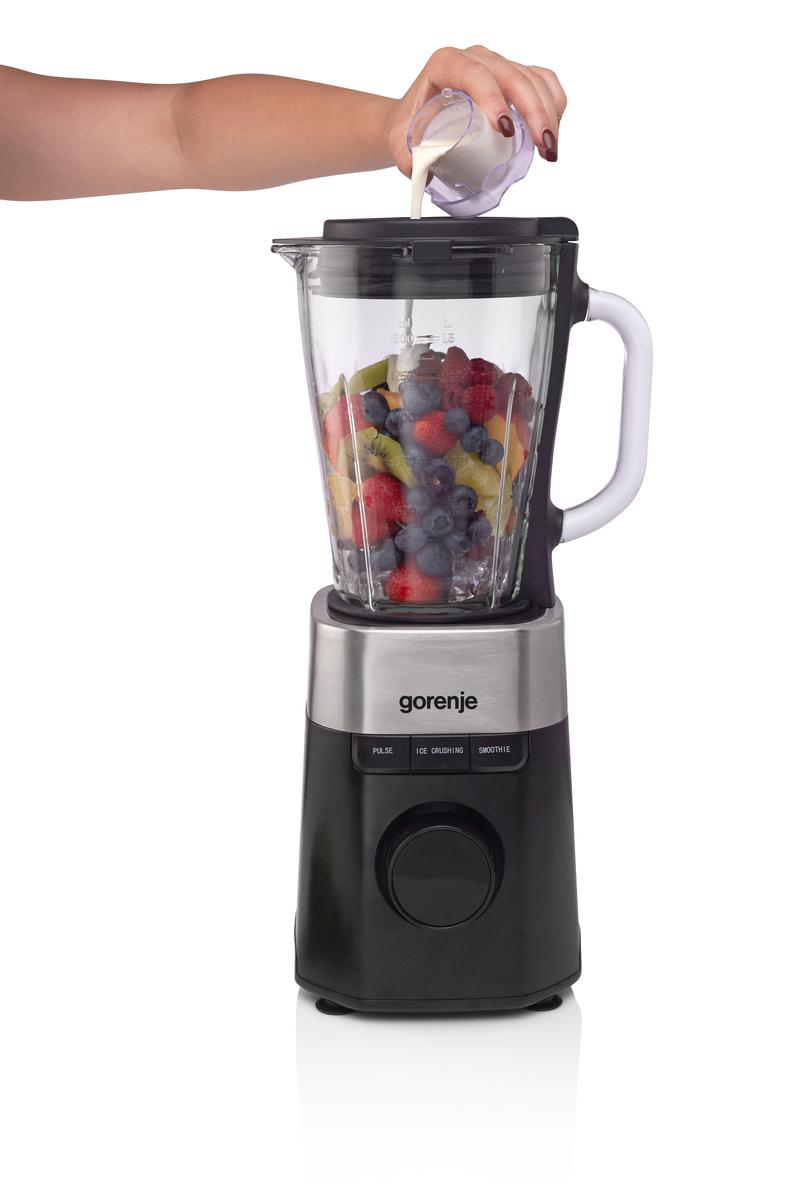 Ein Mixer mit Früchten darin. Eine Hand gießt Milch aus einem kleinen Krug in den Mixer. Der Mixer ist schwarz, silberfarben und aus Glas.