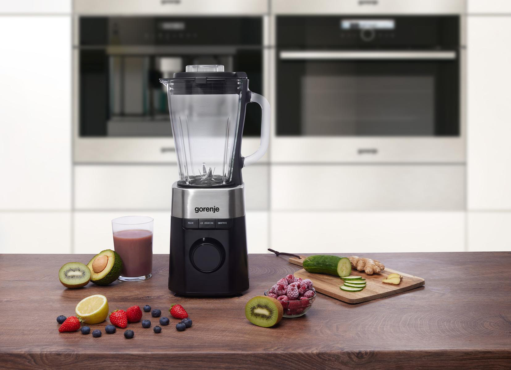 Ein Mixer mit Früchten und Gemüse auf einem Holztisch. Enthält Kiwi, Beeren, Zitrone, Gurke und Ingwer.