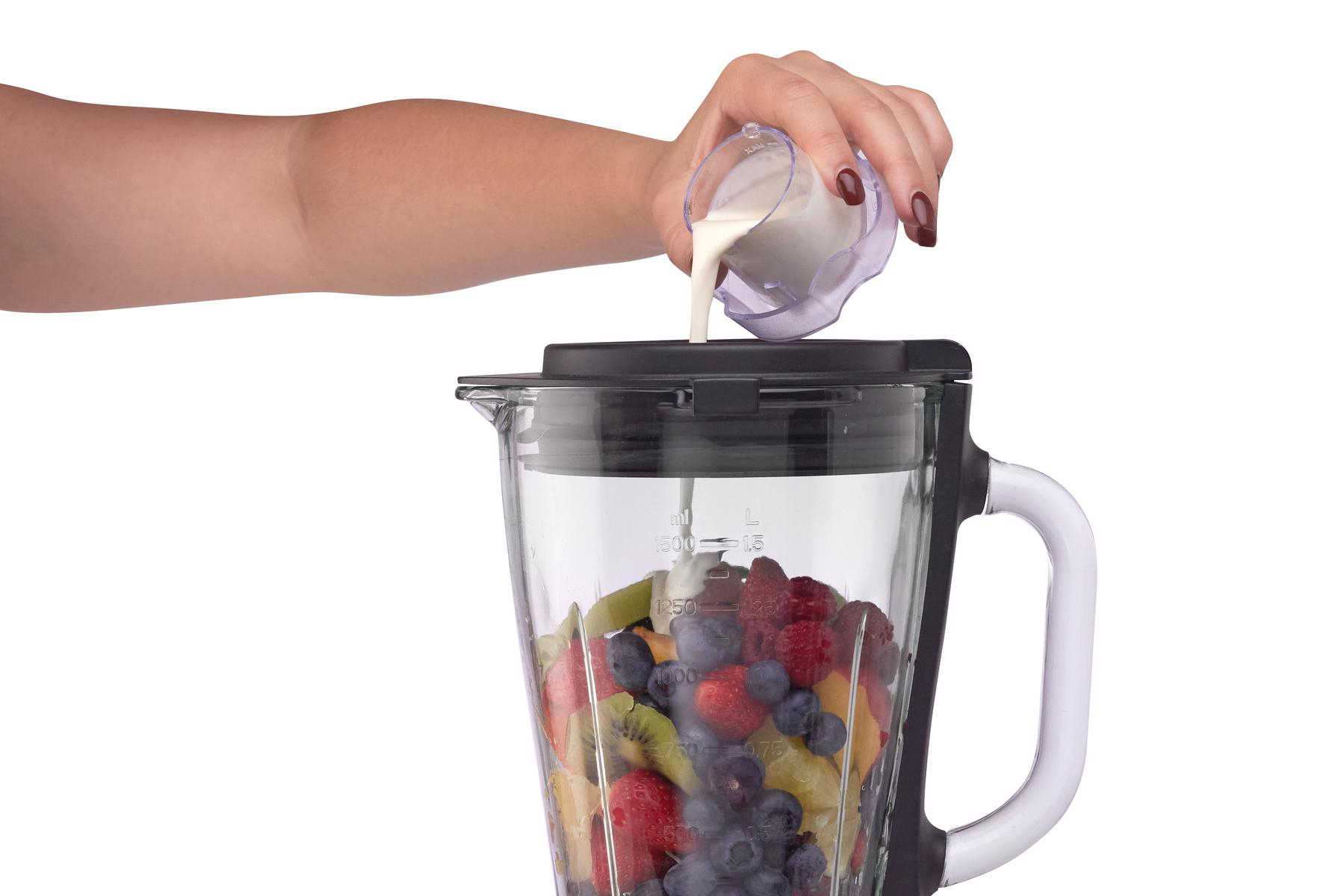Eine Hand gießt Milch in einen Mixer, der mit Früchten gefüllt ist. Rote, grüne und blaue Früchte sind sichtbar. Der Mixer ist klar.