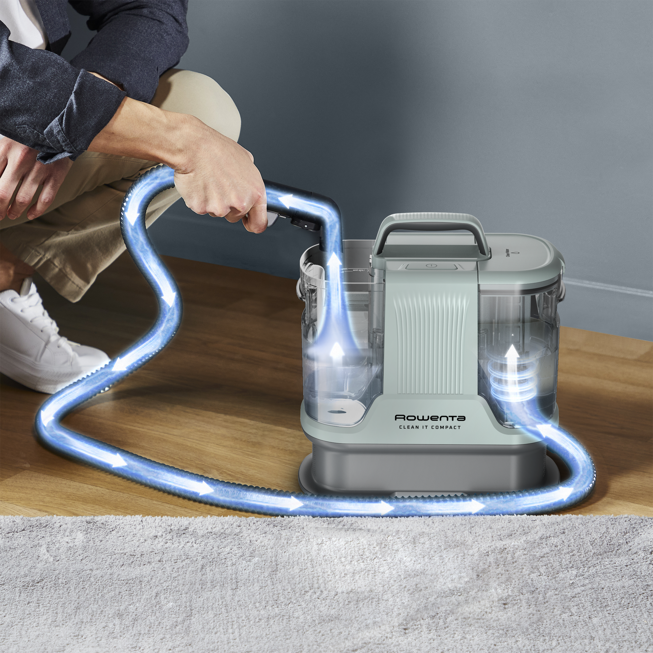 Egy személy egy Rowenta Clean It Compact porszívót használ. A kék nyilak a légáramlást mutatják. A porszívó fa padlón és egy fehér szőnyegen van.