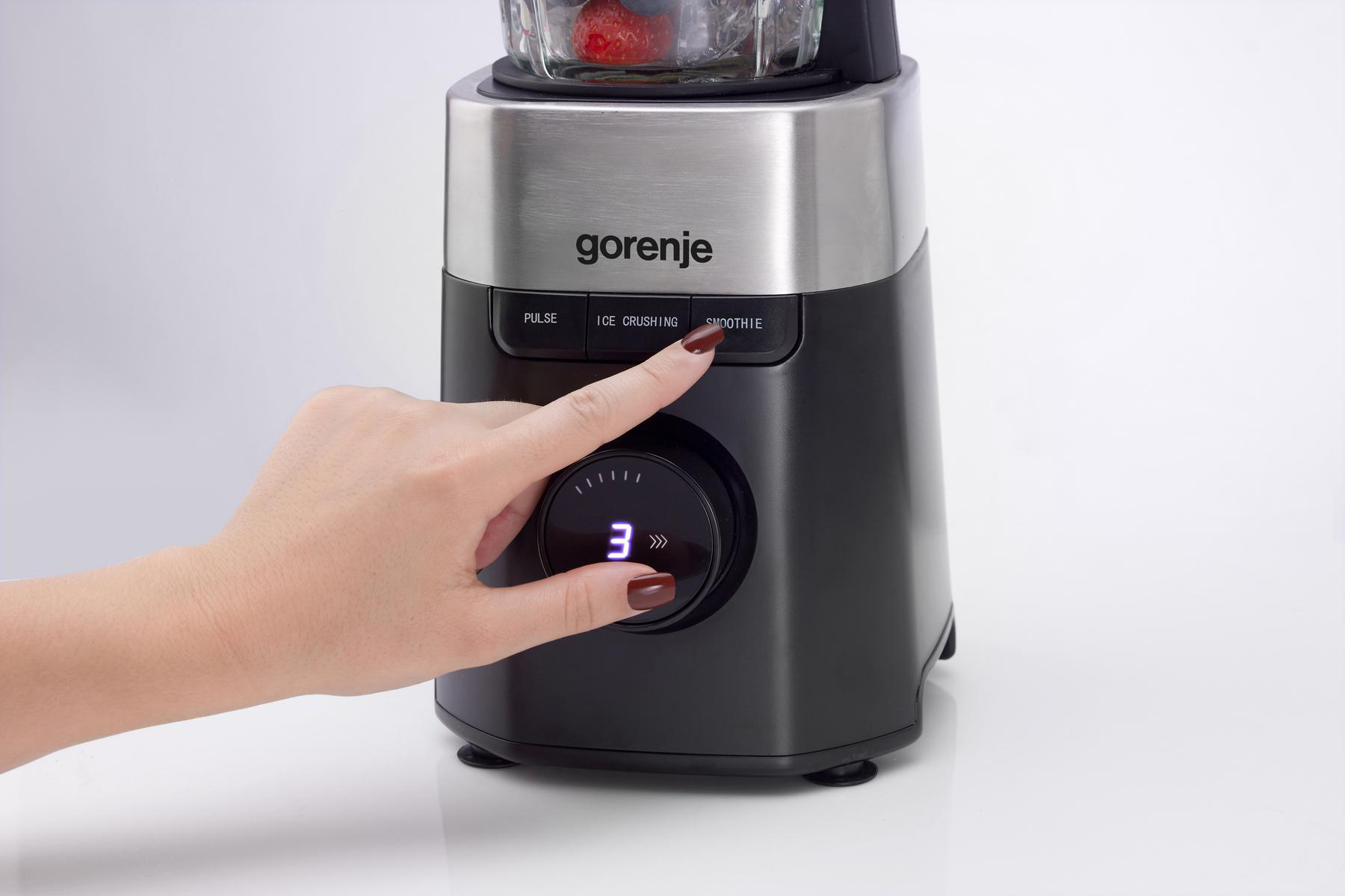 Eine Hand drückt die Taste 'Smoothie' an einem schwarzen Gorenje-Mixer. Das Zifferblatt zeigt '3'. Der Mixer hat einen Glasbehälter mit Früchten.