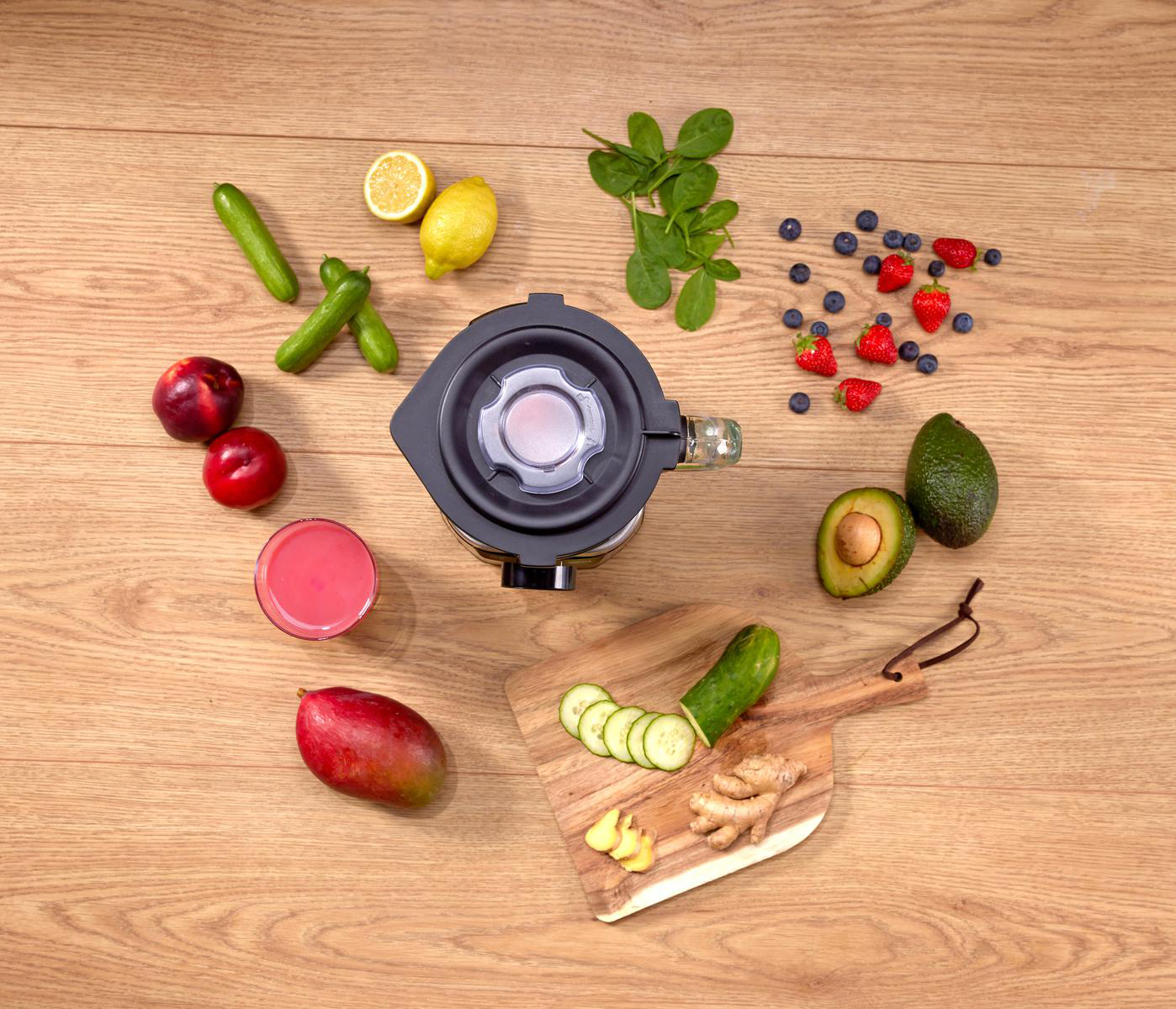 Mixer umgeben von Obst und Gemüse: Gurken, Zitronen, Pfirsiche, Spinat, Beeren, Avocado, Mango und Ingwer auf Holz.