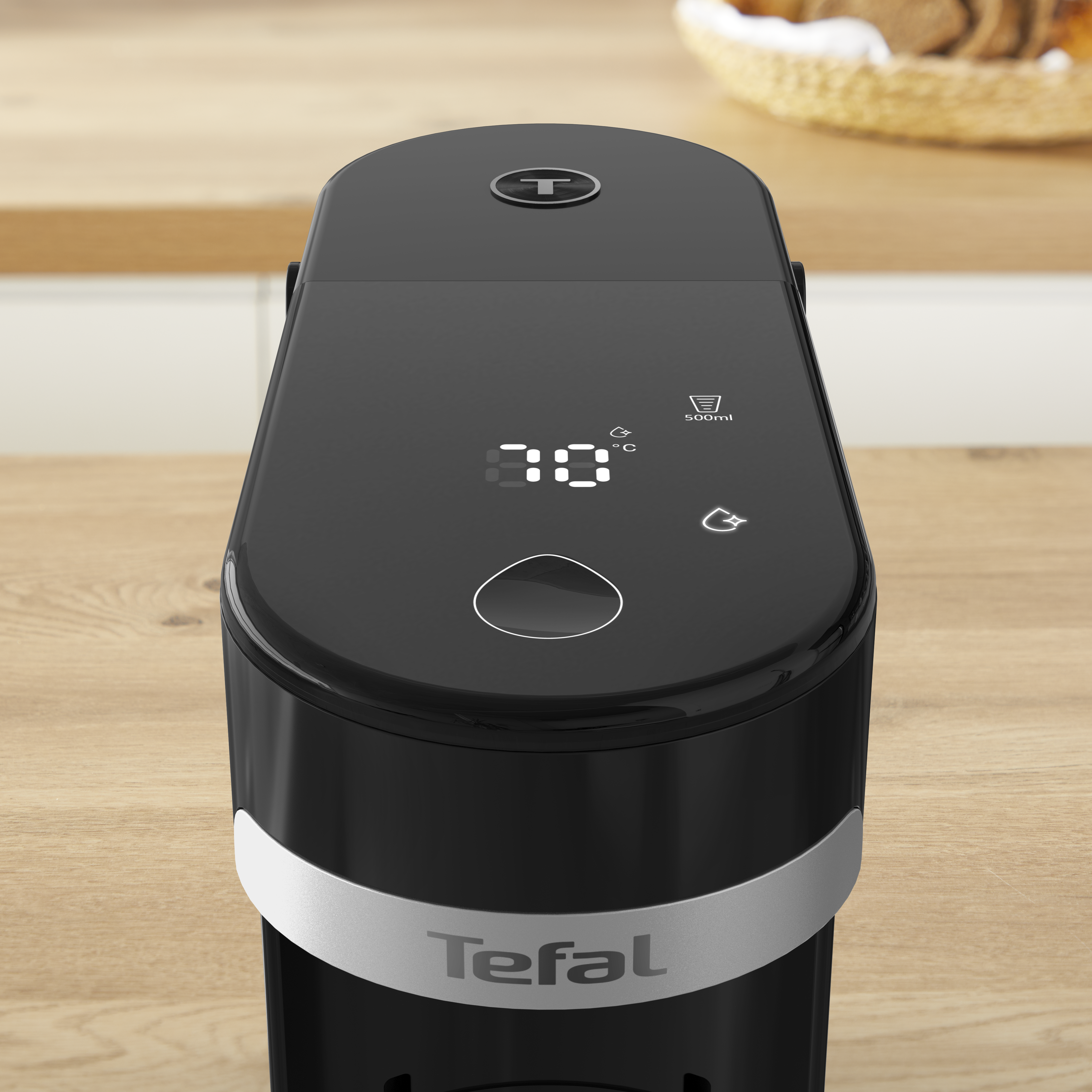 Fekete Tefal vízforraló fa felületen, 70°C-ot mutat. Ezüst sávval és Tefal logóval. A háttérben egy kenyérkosár.