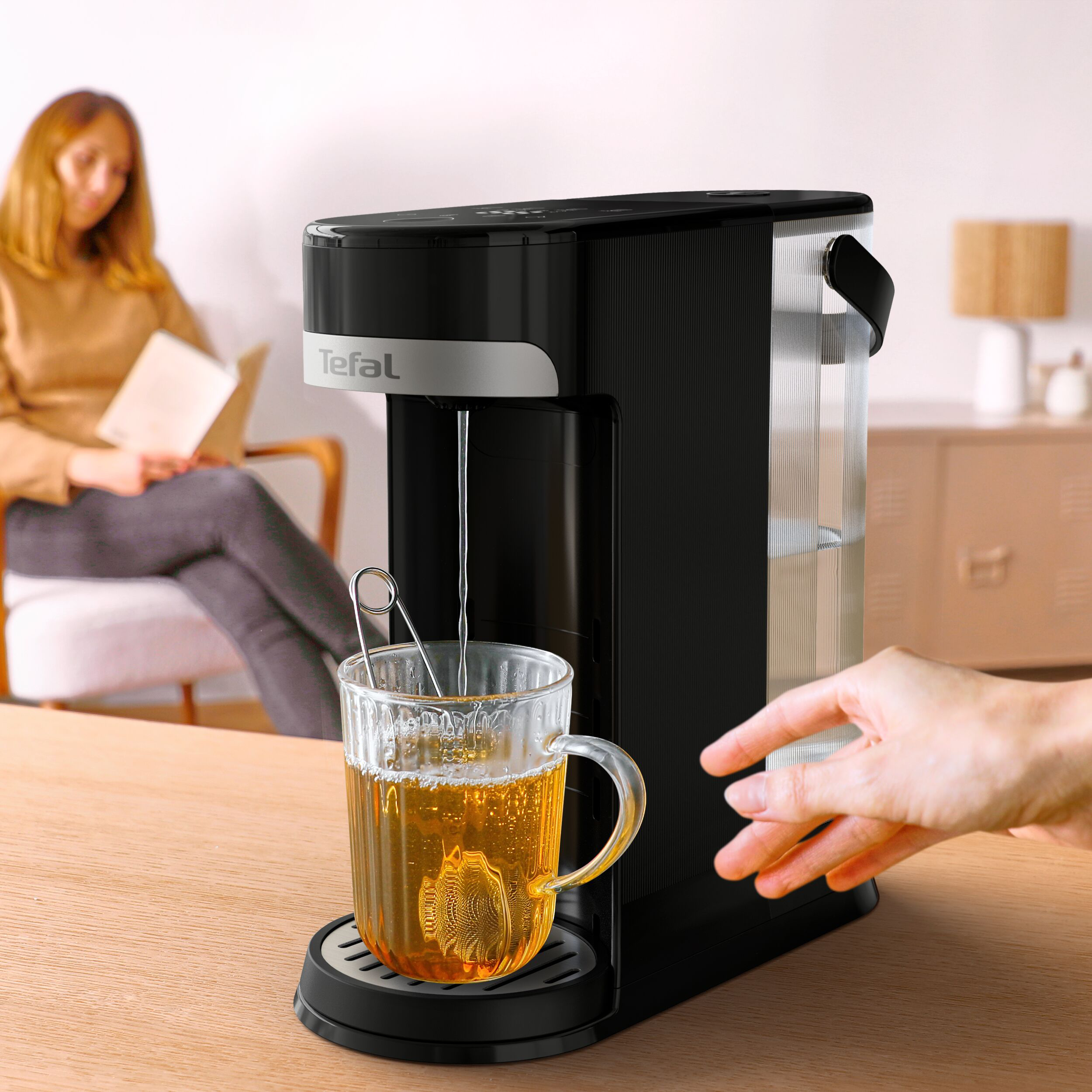 Teafőző üvegpohárral. A háttérben egy személy olvas. Egy kéz a teafőző felé nyúlik.