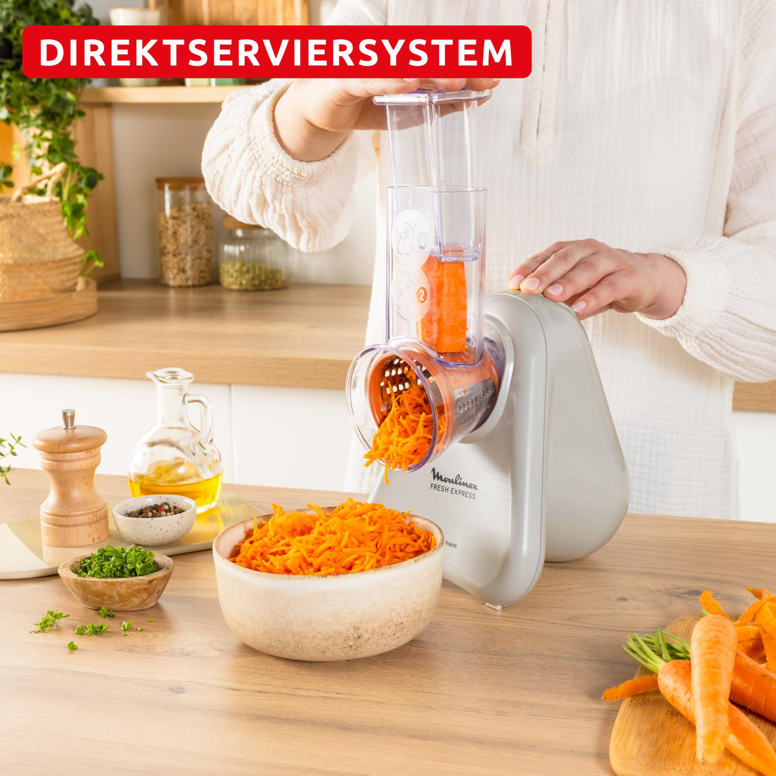 Eine Person raspelt Karotten mit einem Moulinex Fresh Express. Geriebene Karotten fallen in eine Schüssel. Weitere Zutaten sind auf dem Tisch.