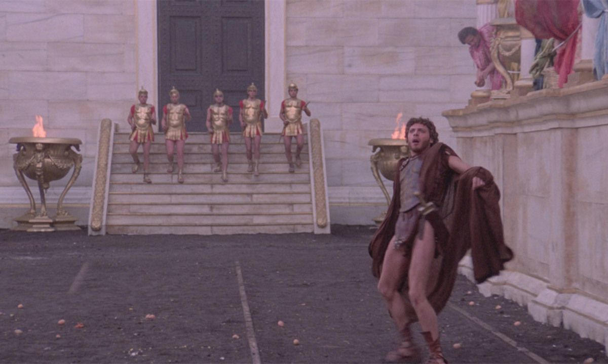 Ein Mann in einer Toga sieht überrascht aus. Soldaten in Rüstungen steigen eine Treppe hinunter. Links eine brennende Urne, Figuren auf einem Balkon.