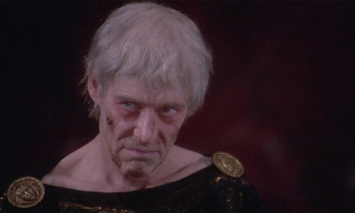Ein älterer Mann mit grauem Haar und ernstem Blick. Er hat Gesichtsverletzungen und trägt eine dunkle Robe mit goldenen Akzenten.
