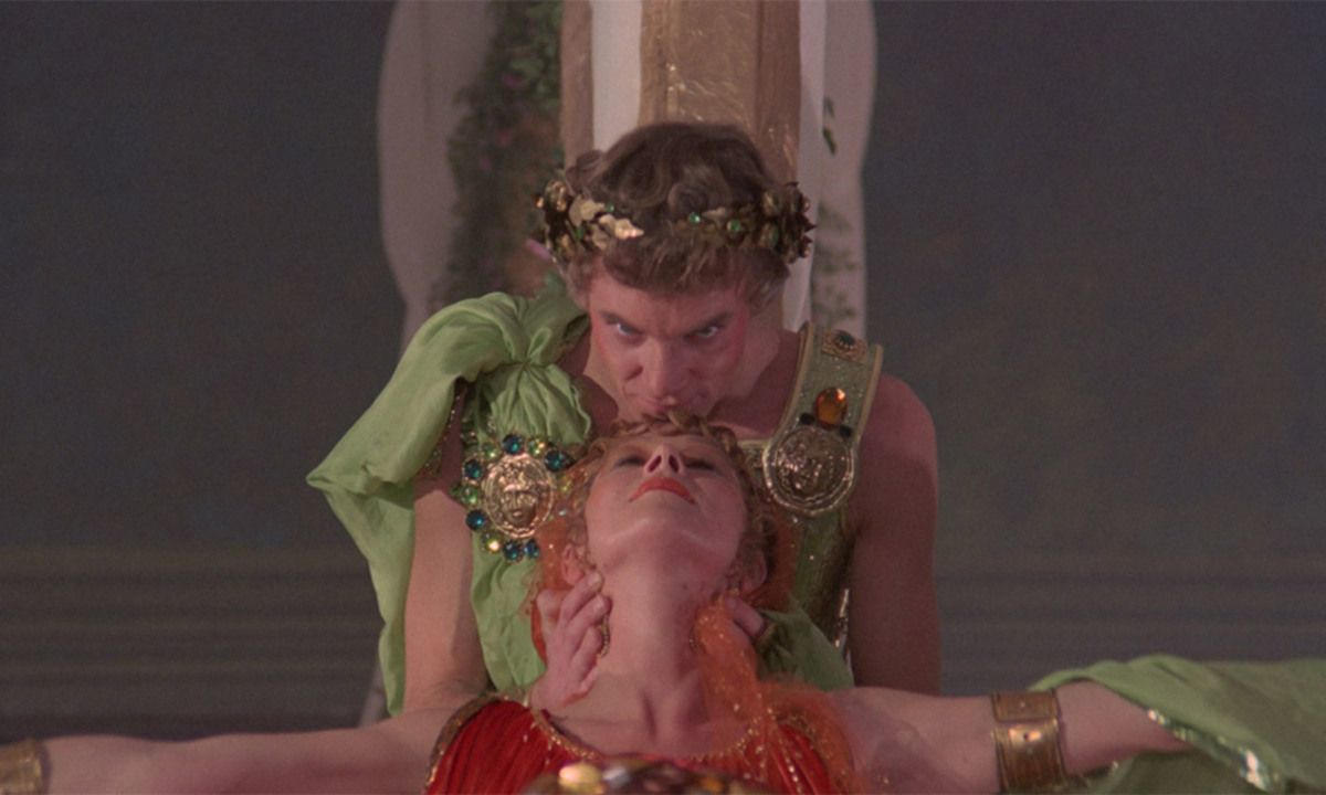 Ein Mann küsst die Stirn einer Frau. Beide tragen kunstvolle Kleidung, er eine Krone und sie rotes Haar, in einer theatralischen Umgebung.
