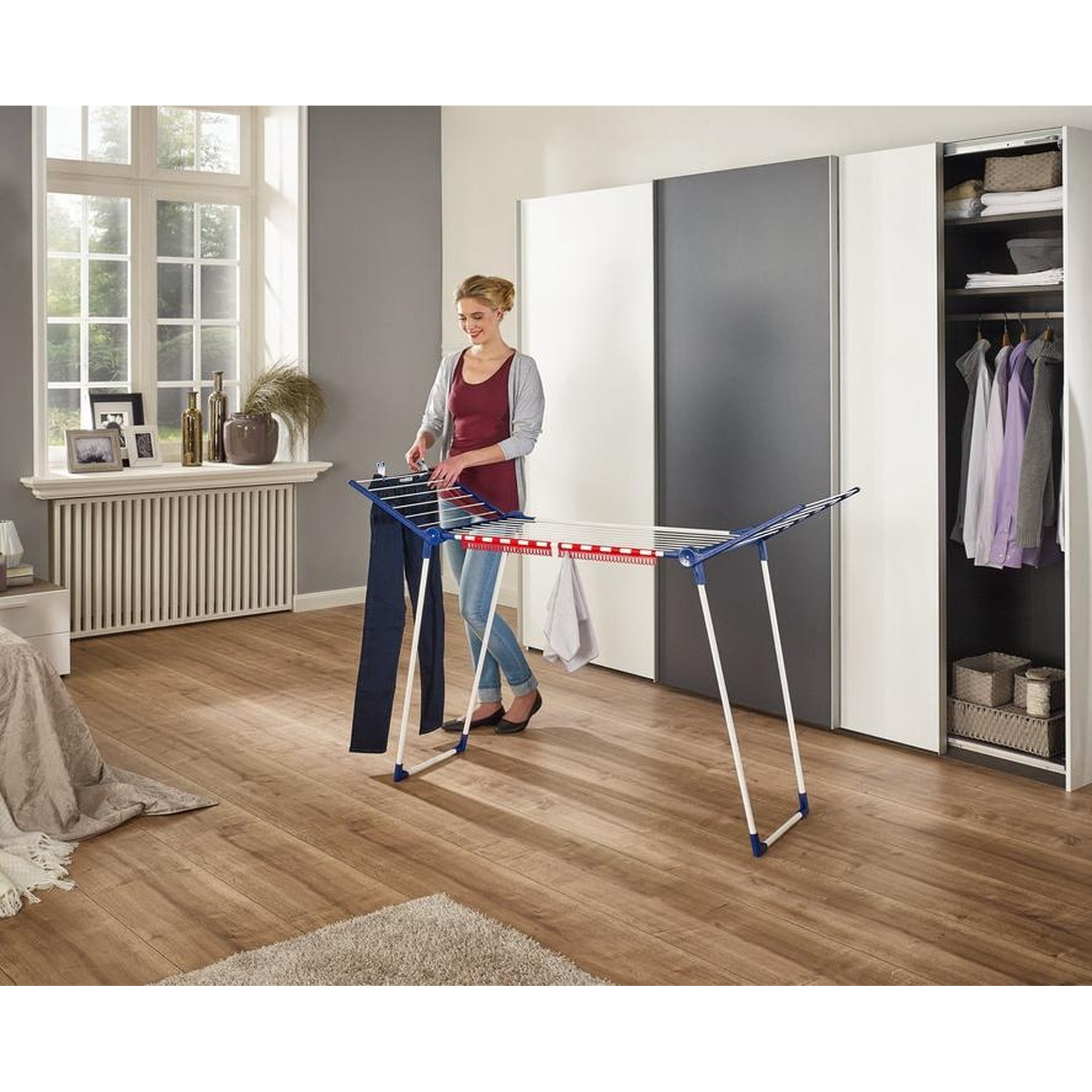 Une femme accroche du linge sur un étendoir dans une chambre. Une fenêtre, un placard et un tapis sont également visibles.