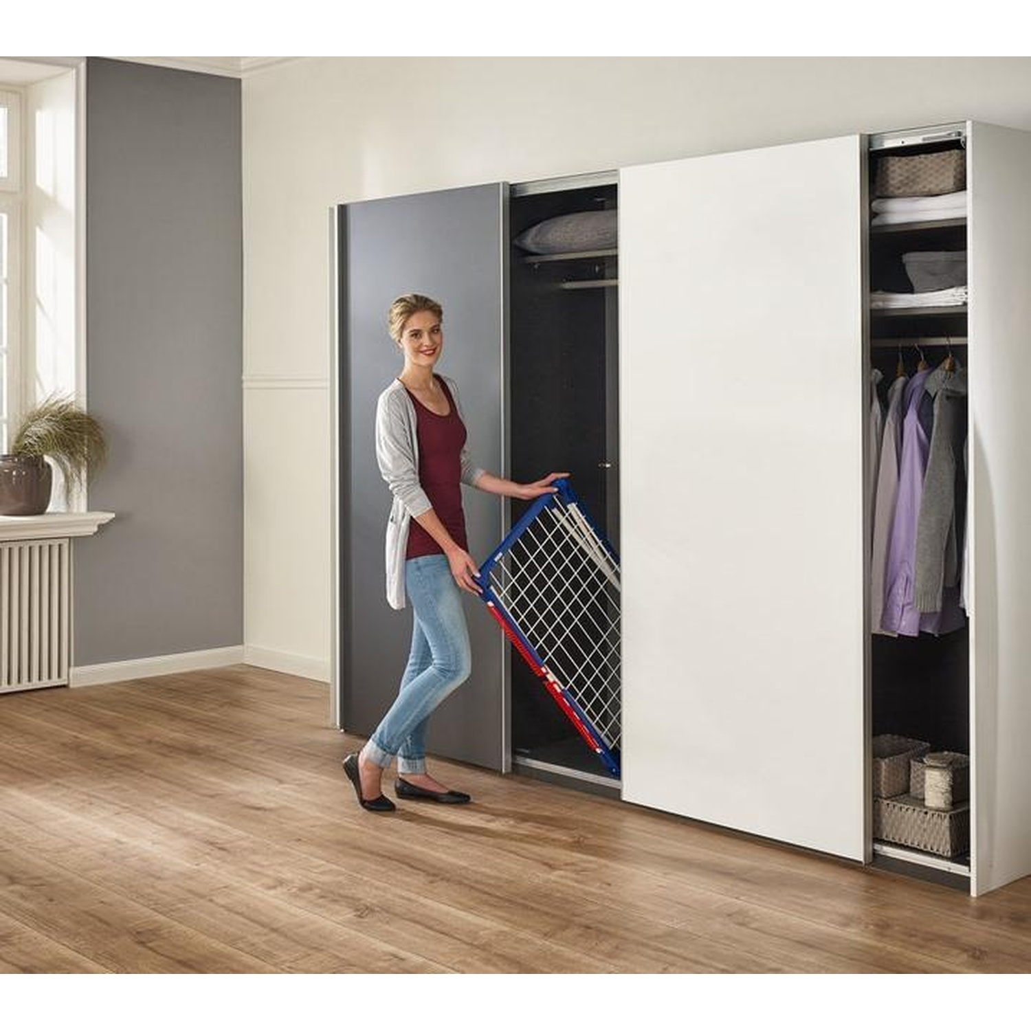 Femme dans une pièce avec une armoire. Elle tient un étendoir bleu et rouge. L'armoire est ouverte, révélant des vêtements et des étagères.