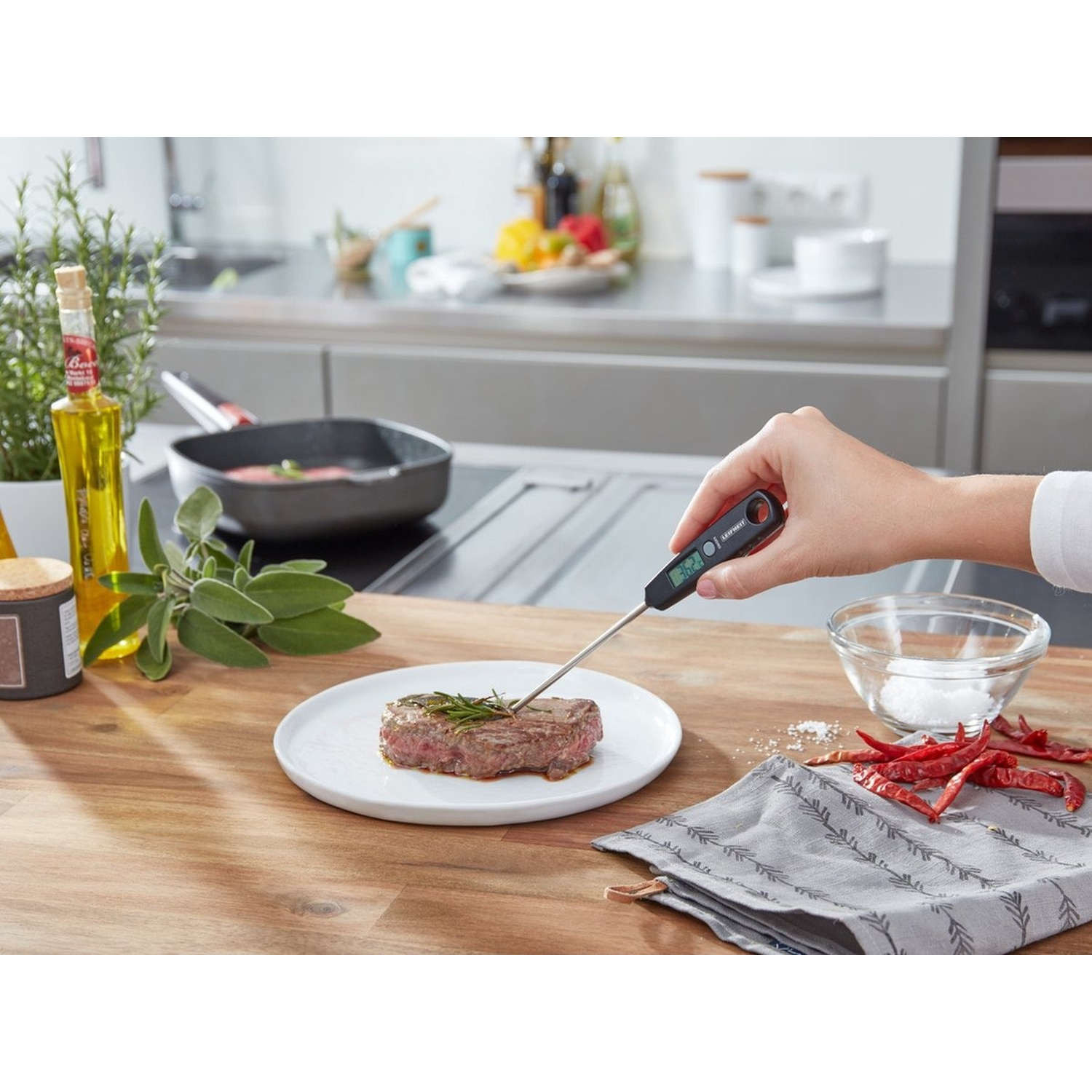 A person uses a thermometer to check the temperature of a steak on a plate. Various ingredients surround it.