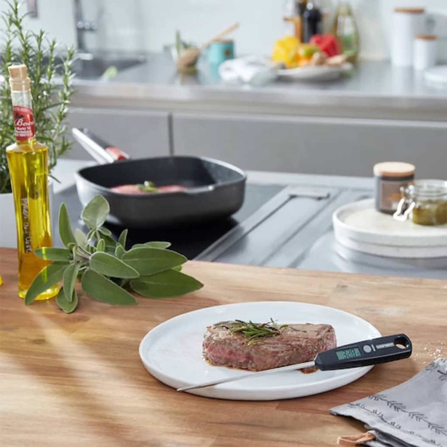 Steak on a white plate with a thermometer. Pan with steak in the background. Olive oil and herbs are on the side. Kitchen setting.