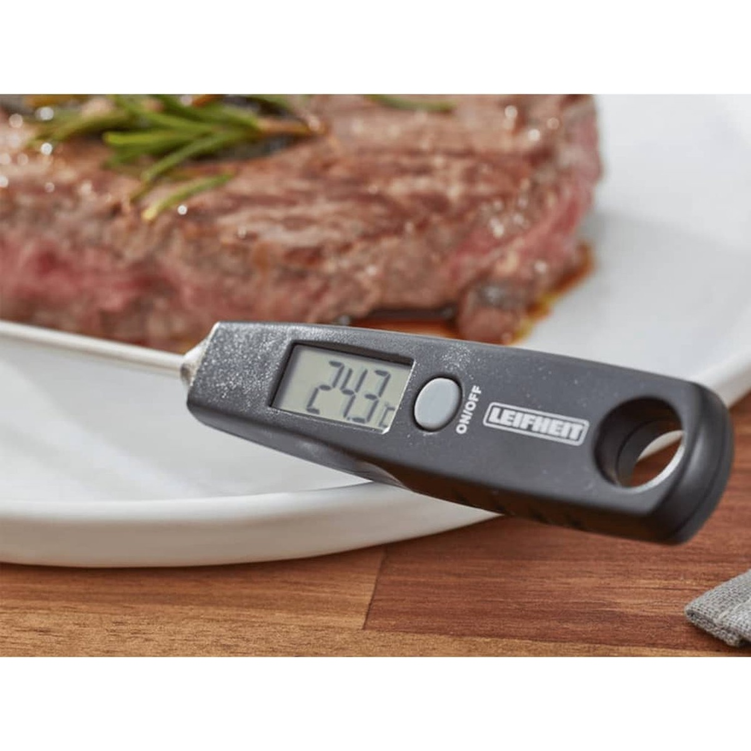 A digital thermometer is inserted into a steak on a white plate. The thermometer shows 24.3 degrees. Rosemary is on the steak.