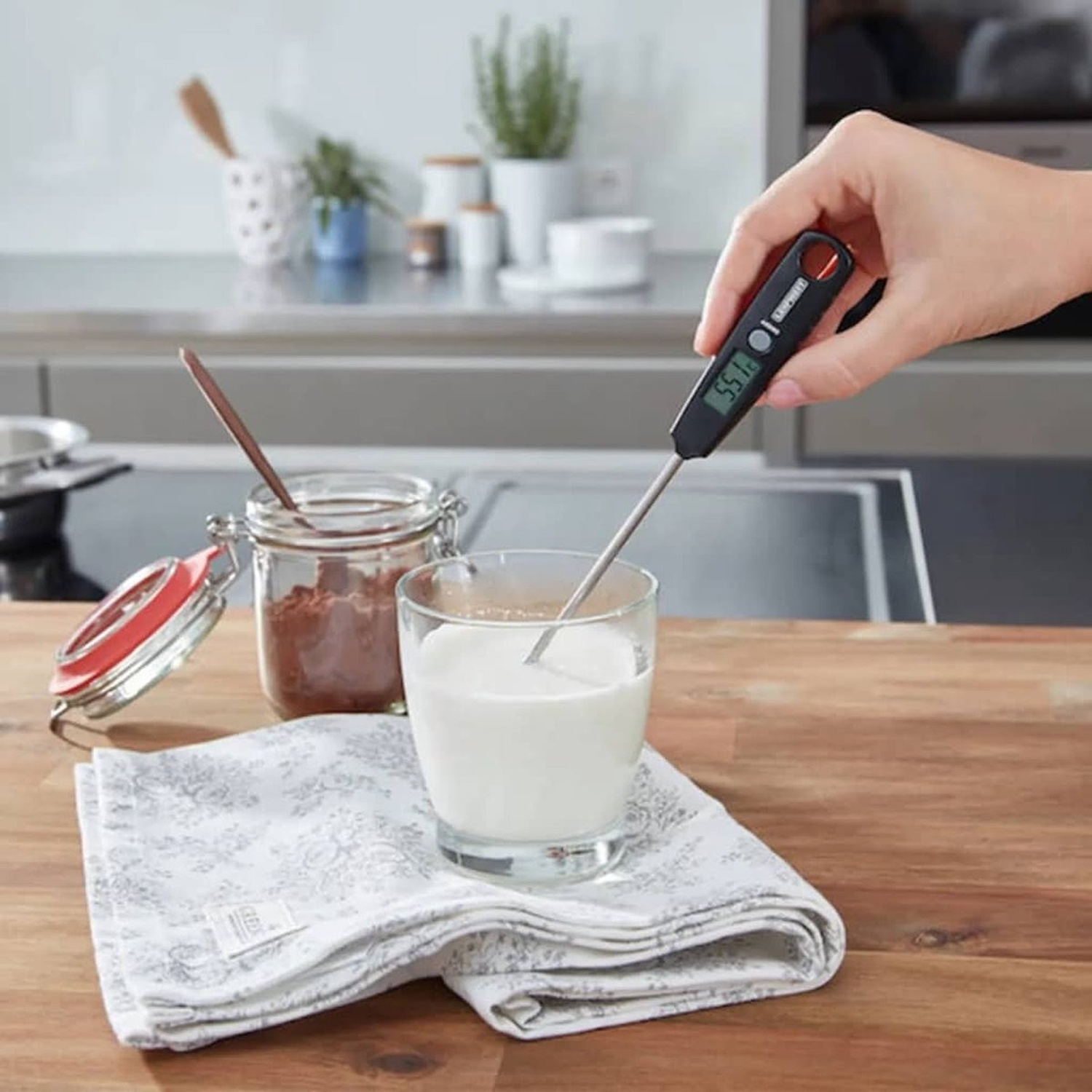 A hand holds a thermometer in a glass of milk. Beside is a jar with cocoa powder and a patterned cloth.