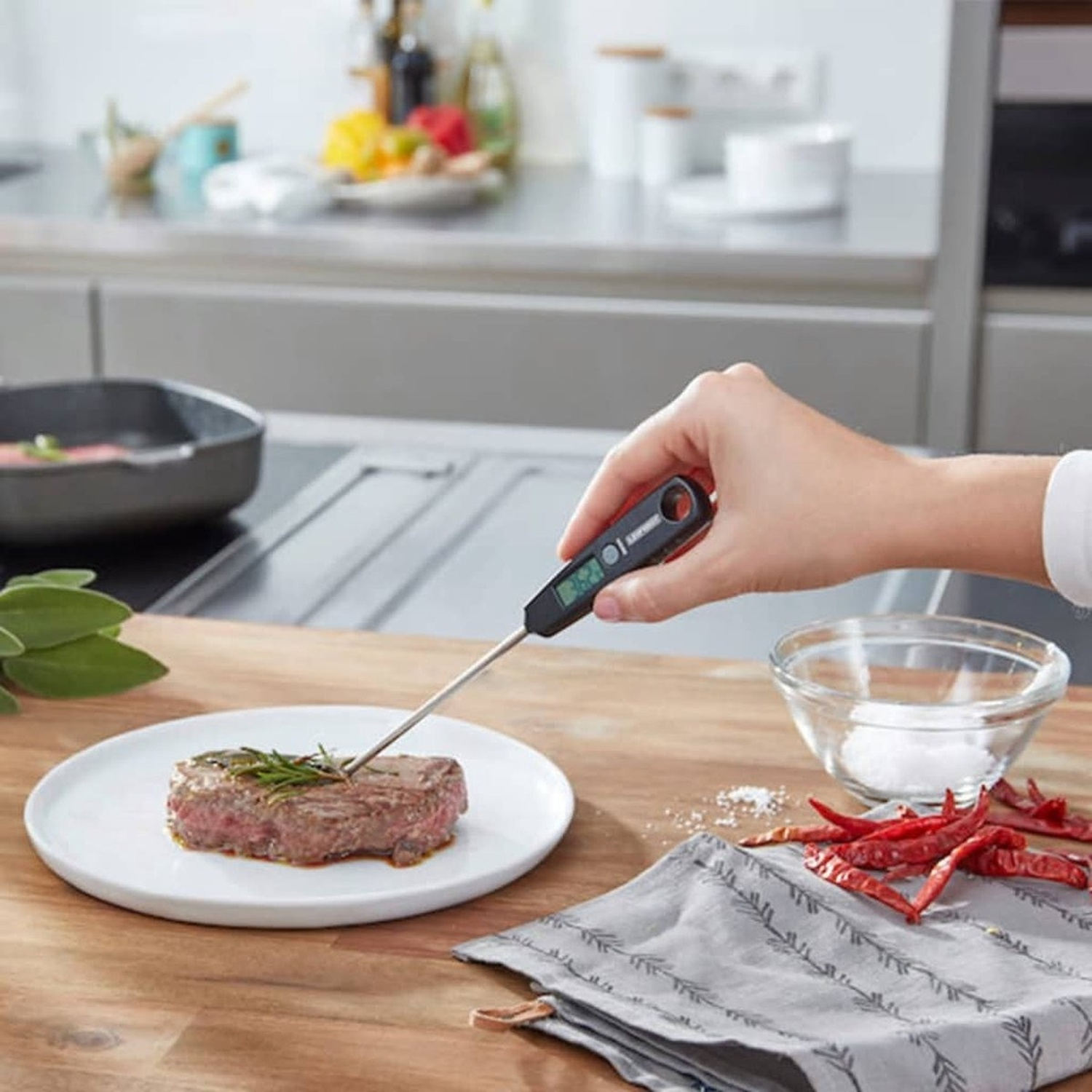 A hand inserts a meat thermometer into a steak on a plate. Spices, salt, and chili peppers are nearby on a wooden surface.