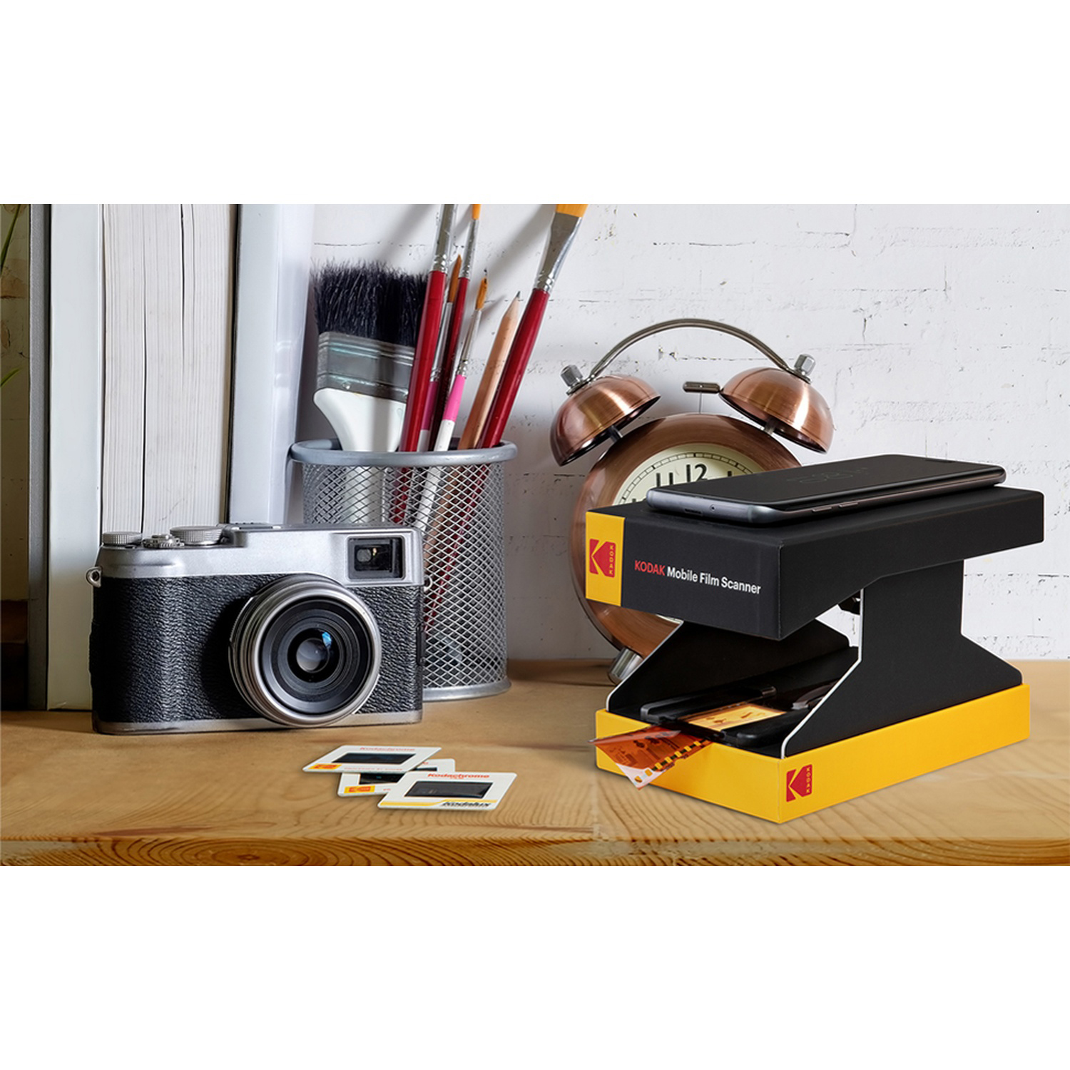 Una fotocamera, uno scanner per pellicole e diapositive su una scrivania di legno. Una sveglia e dei pennelli sullo sfondo.