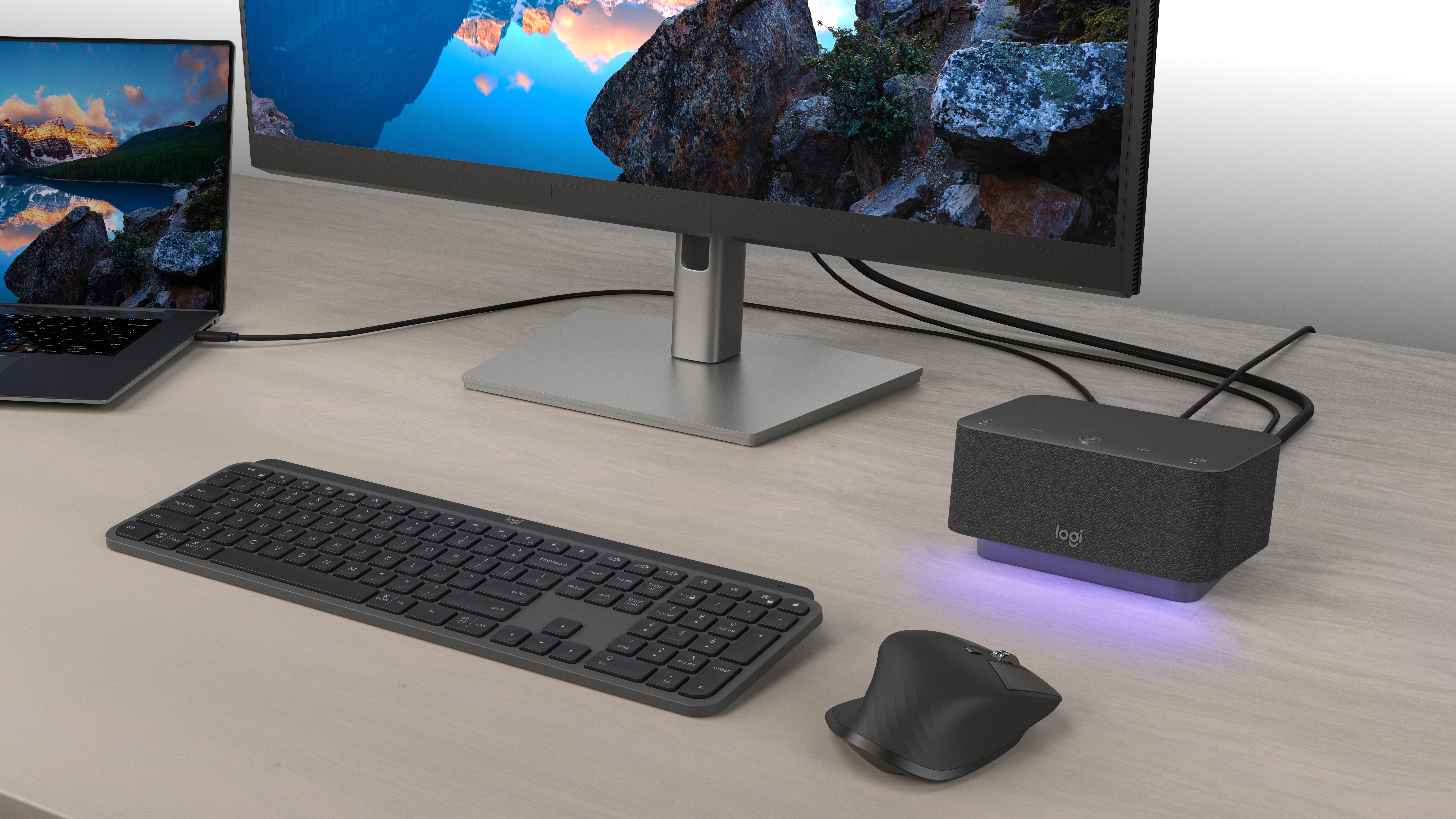 Ein Schreibtisch-Setup mit Monitor, Laptop, Tastatur, Maus und Lautsprecher. Der Monitor zeigt eine Berglandschaft. Alle Gegenstände sind grau.
