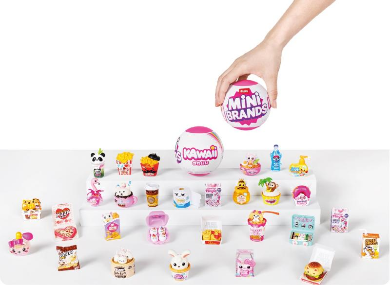 Eine Hand hält einen Mini Brands Ball. Miniaturen von Lebensmitteln werden auf einer weißen Oberfläche ausgestellt, darunter Pommes, Pizza und ein Burger.