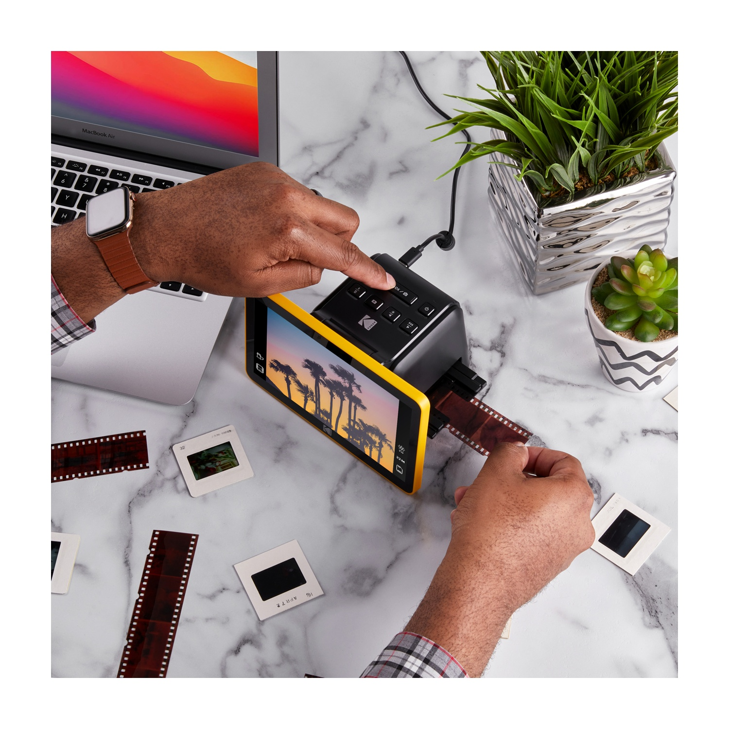 Una persona scansiona negativi e diapositive con uno scanner. Sono visibili un laptop e delle piante. Lo scanner ha una cornice gialla.