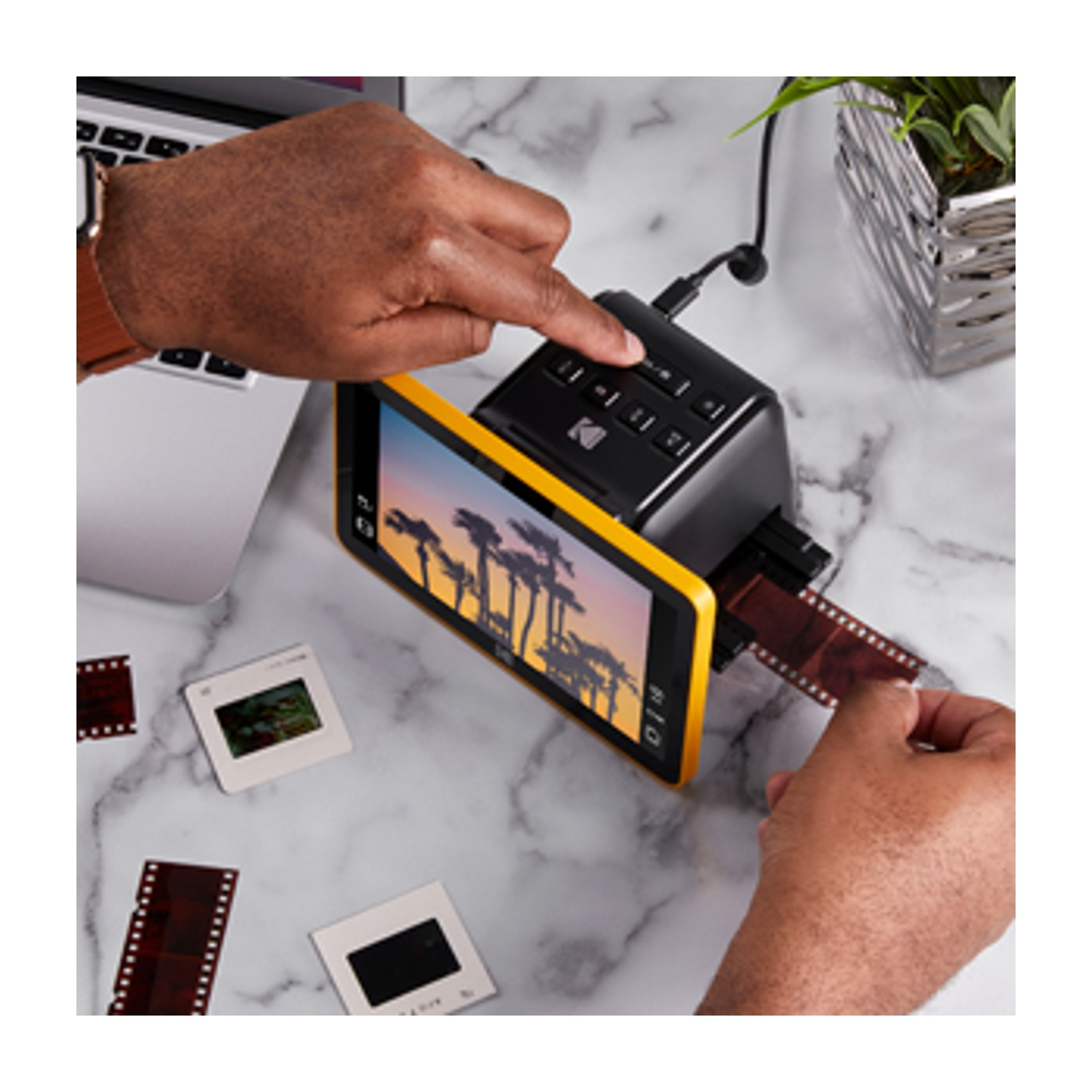 Mani che utilizzano uno scanner per film. Lo scanner ha una cornice gialla. Strisce di film e diapositive sono su una superficie di marmo.