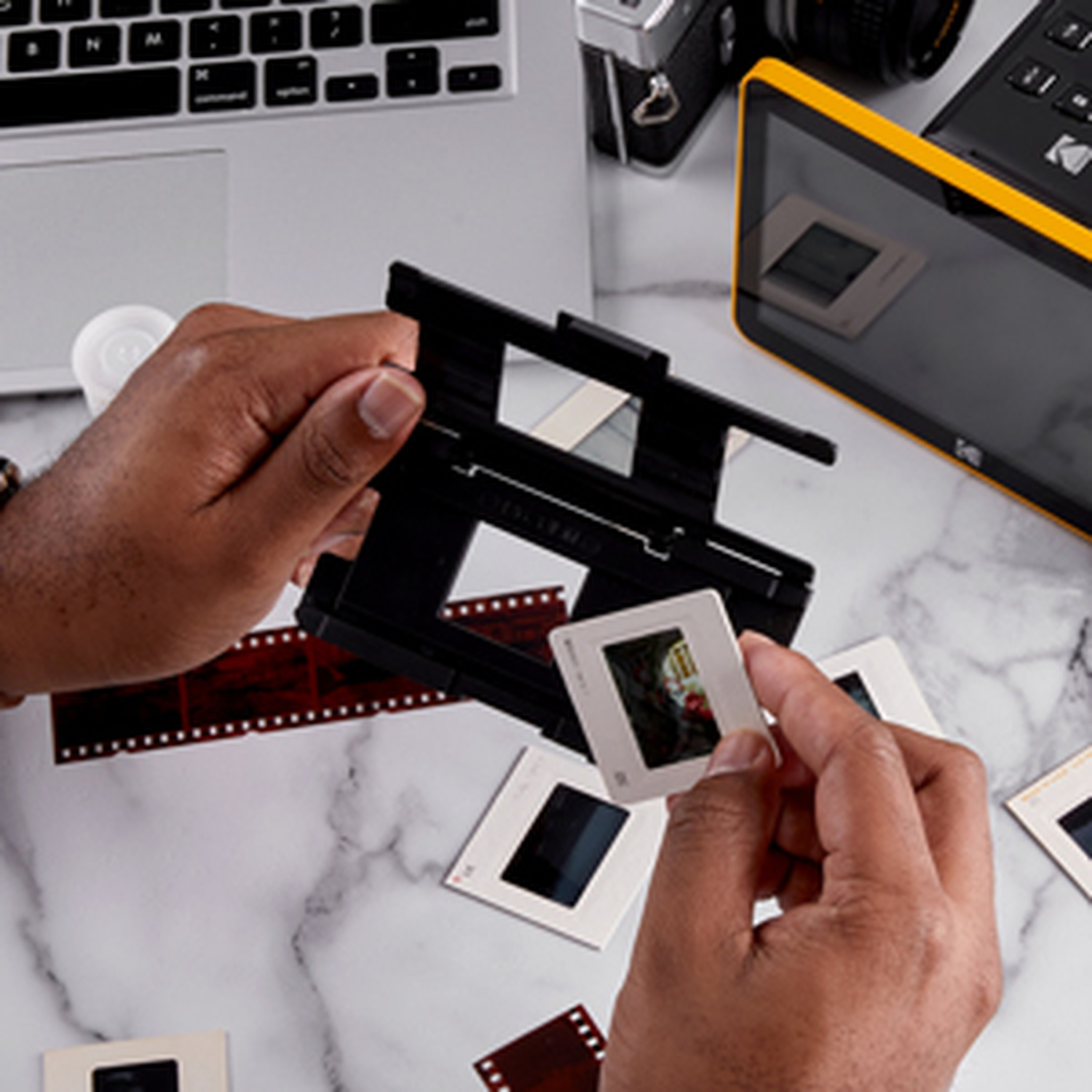 Mani che tengono una diapositiva e un supporto nero. Strisce di pellicola e diapositive su un tavolo di marmo con laptop, fotocamera e scanner giallo.