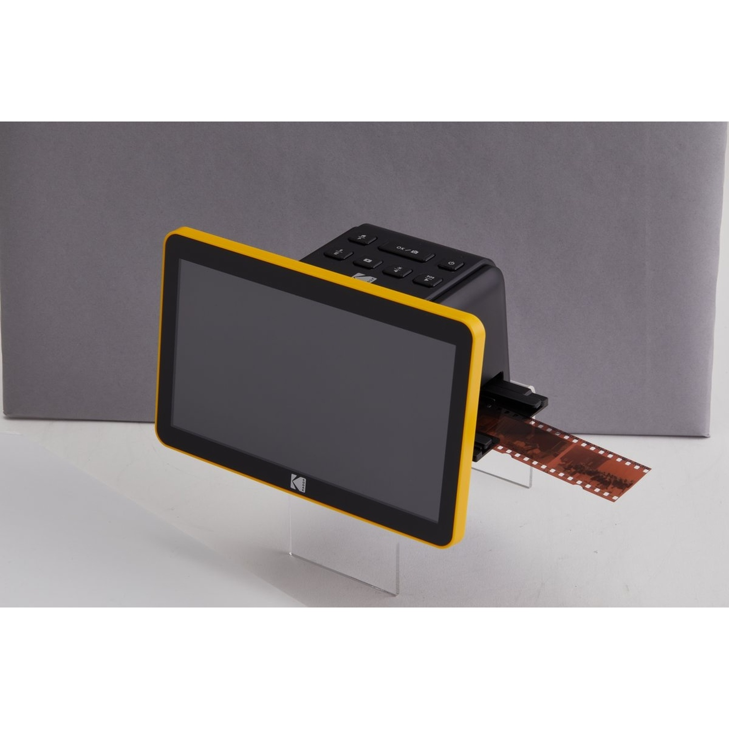 Uno scanner per pellicole Kodak con schermo con cornice gialla, con una striscia di pellicola inserita. Si trova su un supporto trasparente su sfondo grigio.