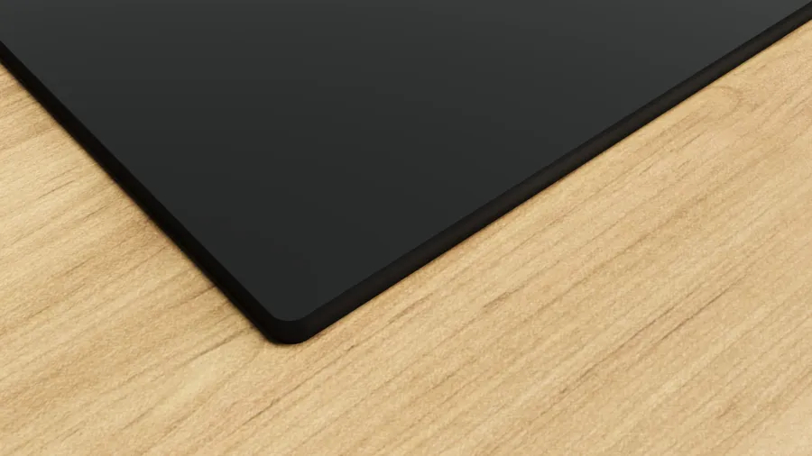 Un objeto negro y rectangular se encuentra en una superficie de madera clara, en la esquina. El objeto tiene una esquina redondeada.