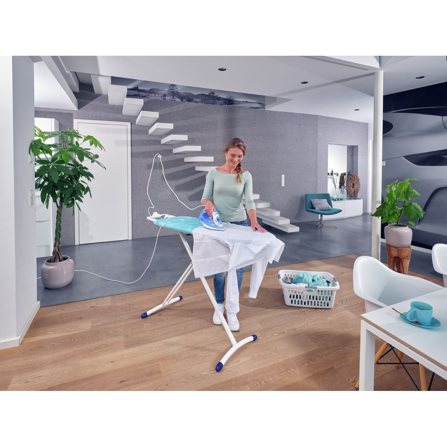 Une femme repasse une chemise blanche sur une planche à repasser. Un panier à linge et un intérieur moderne l'entourent, avec des escaliers et des plantes.