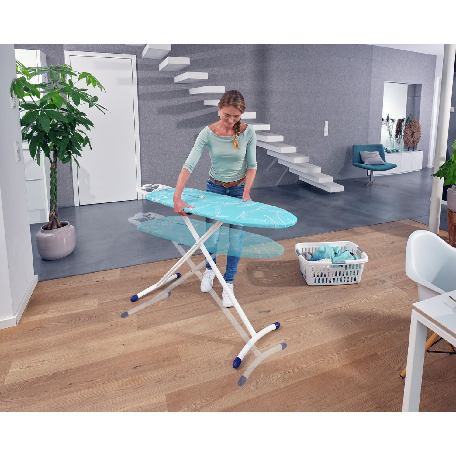 Femme installant une planche à repasser dans une pièce. Panier à linge et plante en arrière-plan. Escalier et chaise visibles.