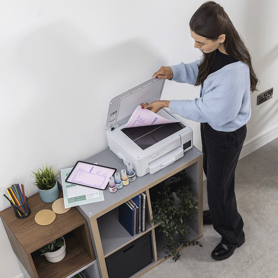 Kobieta wkłada różową fakturę do białej drukarki. Tablet, butelki z tuszem i rośliny znajdują się na półce. Ma na sobie niebieski sweter.