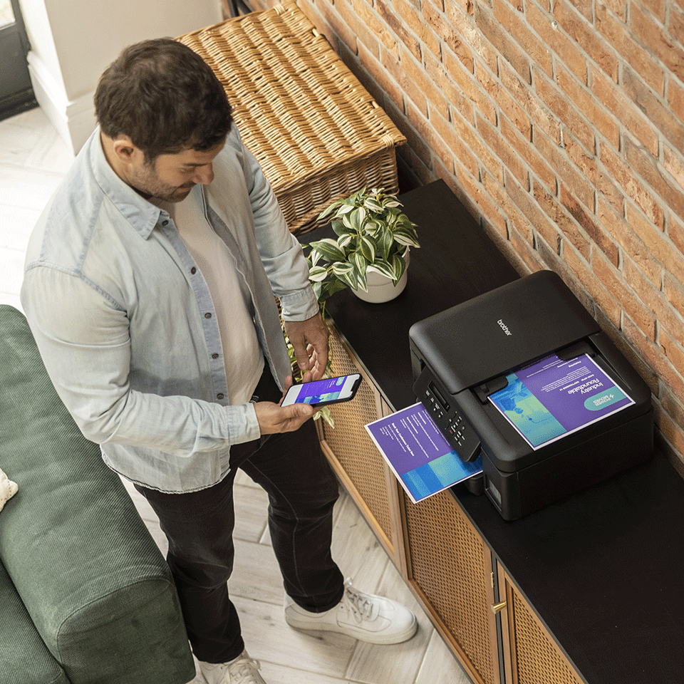 Mężczyzna używa telefonu do drukowania z drukarki Brother. Wiklinowy kosz i roślina znajdują się na czarnej szafce na tle ceglanej ściany.