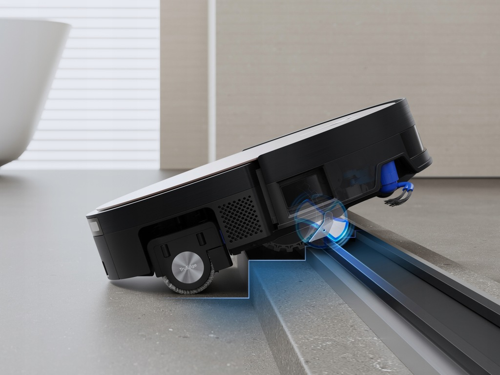 Czarno-biały robot sprzątający czyści ciemnoszarą powierzchnię. Odkurzacz jest pochylony, widać jego koła i elementy czyszczące.