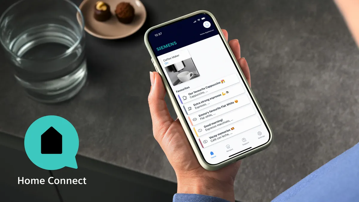 Arka planda bir bardak su ve çikolatalar ile Home Connect uygulamasının gösterildiği bir telefon tutan el.