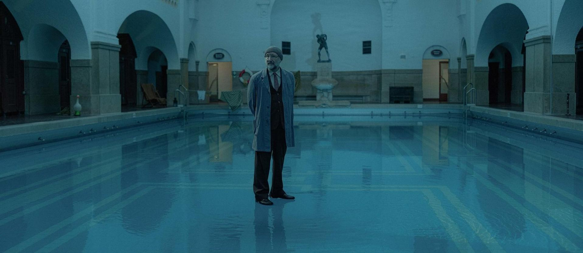 Mann im Mantel steht im Pool. Bögen rahmen die Szene ein. Skulptur und Türen sind im Hintergrund, blaues Wasser spiegelt.