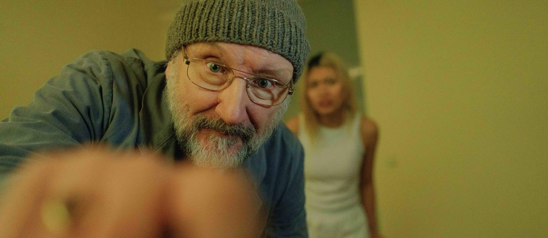 Ein Mann mit Brille und Mütze blickt in die Kamera. Eine Frau ist im Hintergrund unscharf. Die Szene spielt sich drinnen ab.