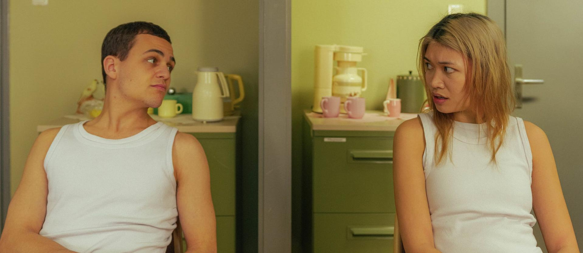 Zwei Personen in weißen Tanktops stehen sich gegenüber. Eine Kaffeemaschine und Tassen stehen auf einem grünen Schrank im Hintergrund.