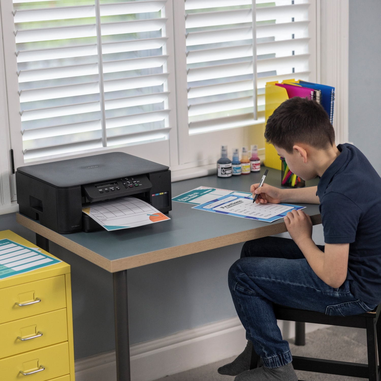 A young boy sits at a desk, writing. A printer is on the desk. A window and a yellow cabinet are in the background.