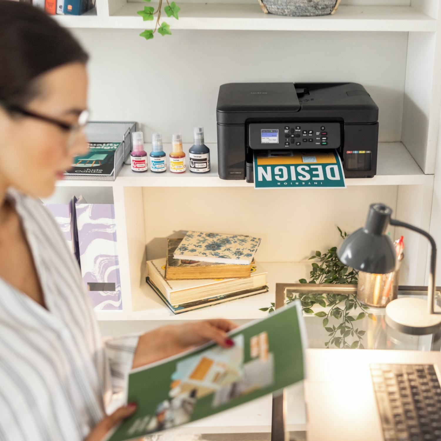 Woman in glasses looks at a magazine. Printer prints 'INTERIOR DESIGN'. Books and ink bottles on shelves. Desk with lamp and laptop.