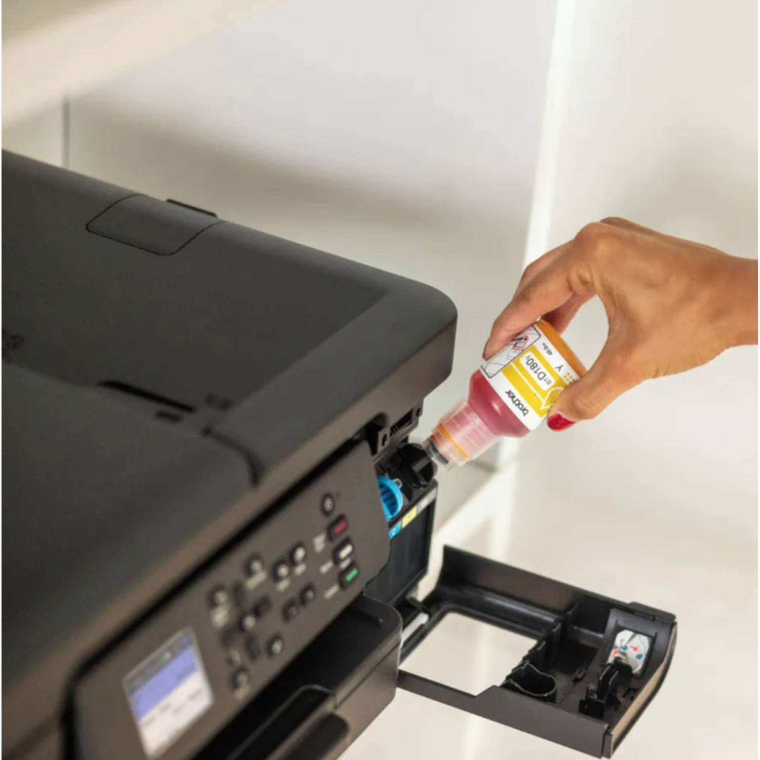 A hand pouring yellow ink into a black printer. The printer is on a white shelf. A bottle with yellow and pink ink.